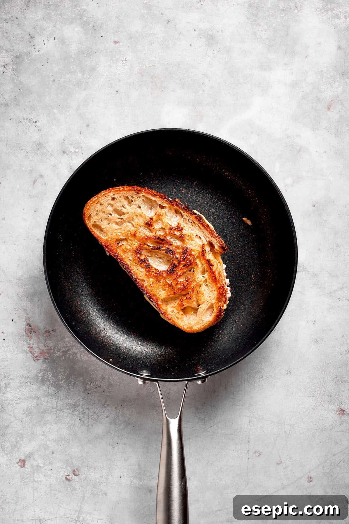 This toasty tuna melt recipe features a blend of creamy tuna salad and melty cheese stuffed between slices of sourdough and pan-fried to a crisp. A tuna melt sandwich is cooking in a non-stick skillet over medium-low heat, with its exterior turning golden brown.