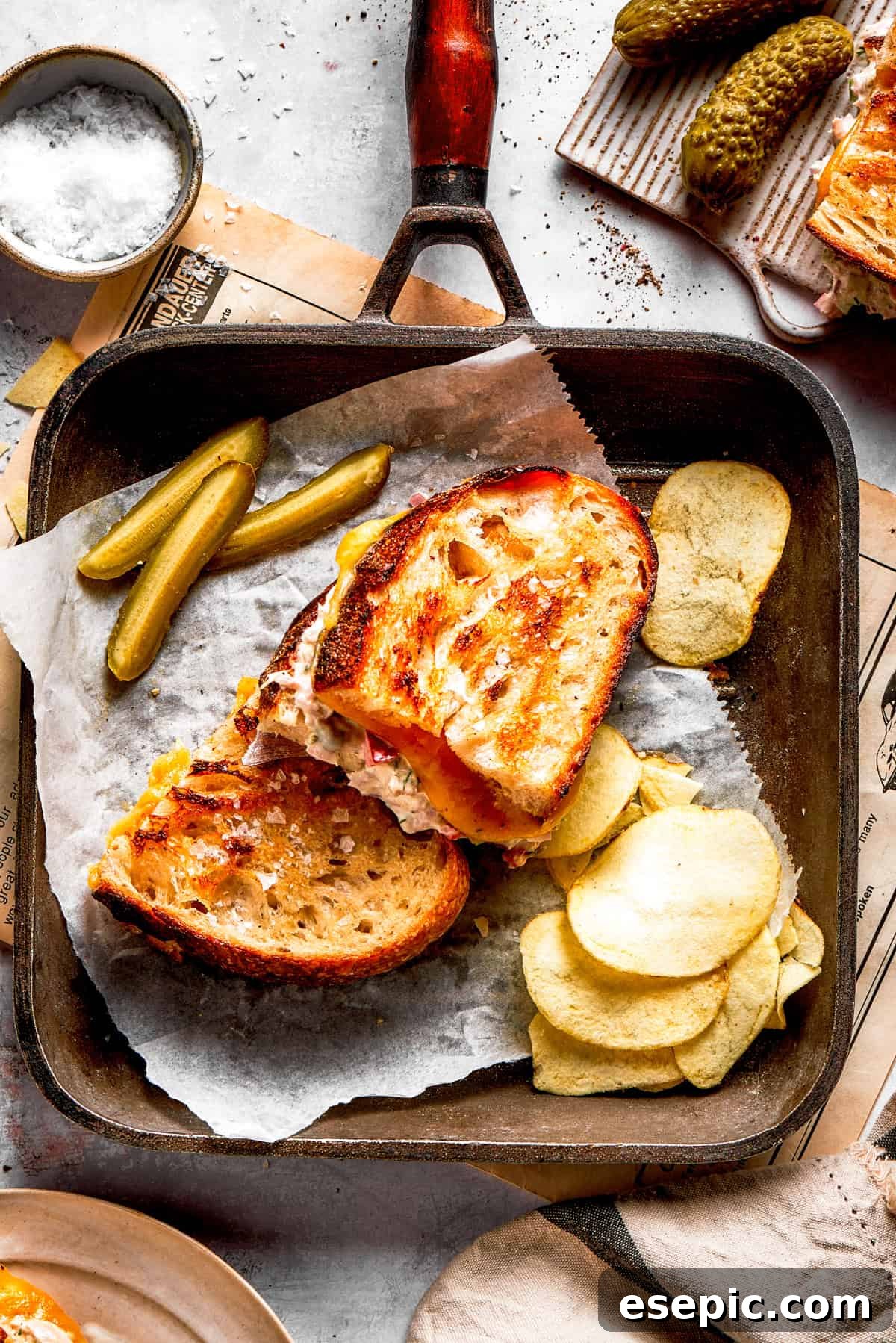 This toasty tuna melt recipe features a blend of creamy tuna salad and melty cheese stuffed between slices of sourdough and pan-fried to a crisp. Two tuna melt sandwiches in a skillet with a pile of potato chips arranged next to them, ready to be served.