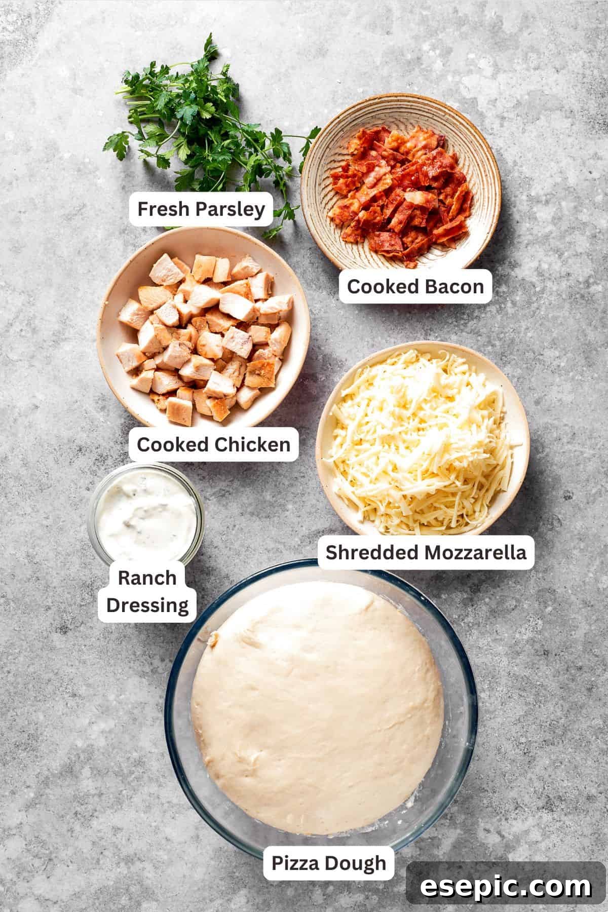 All the essential ingredients for a Chicken Bacon Ranch Pizza laid out on a wooden board, including pizza dough, Ranch dressing, cooked chicken, shredded mozzarella, crispy bacon, and fresh parsley for garnish.
