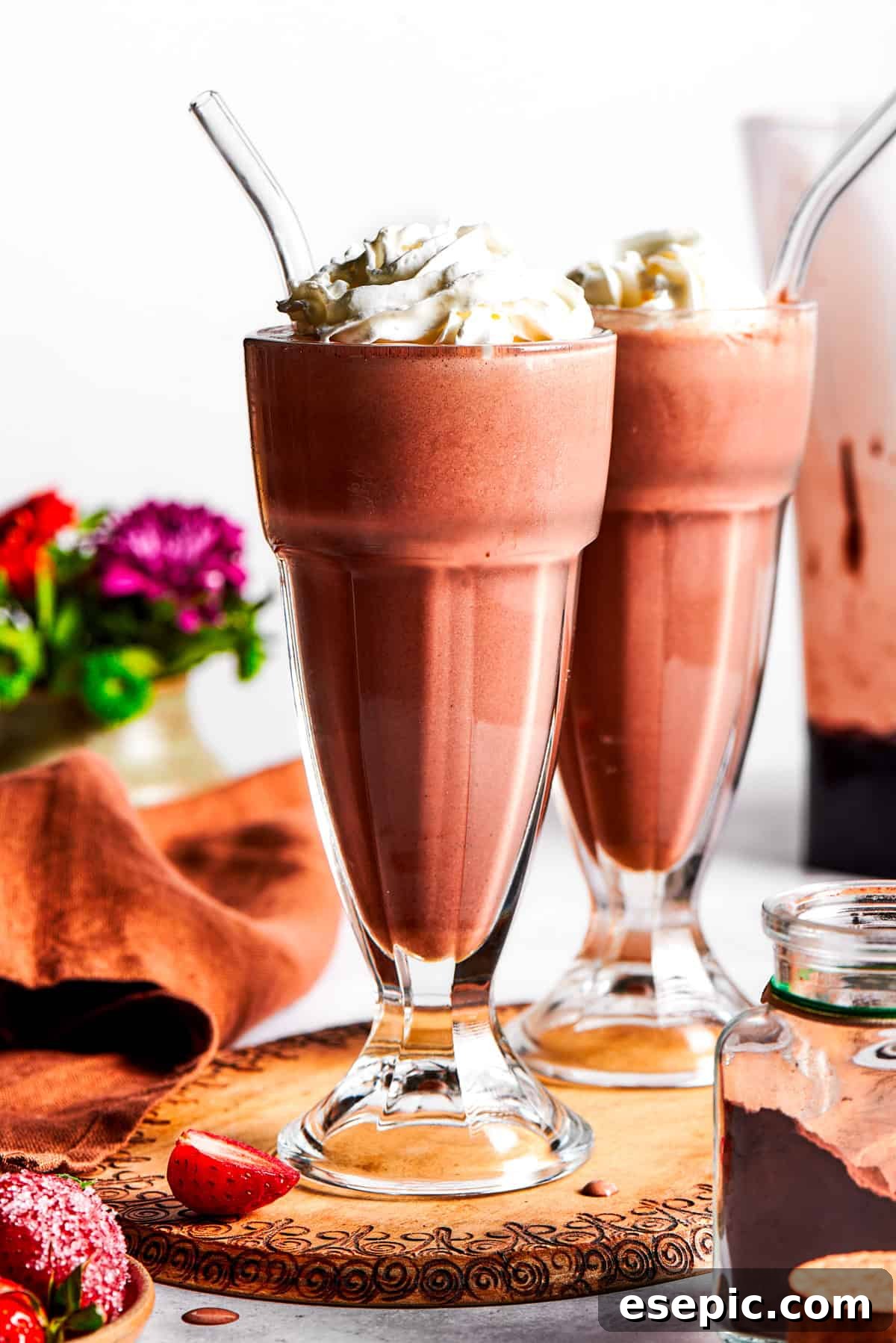 Two glasses of rich chocolate milkshakes topped with generous swirls of whipped cream, a drizzle of chocolate sauce, and fresh strawberry slices.