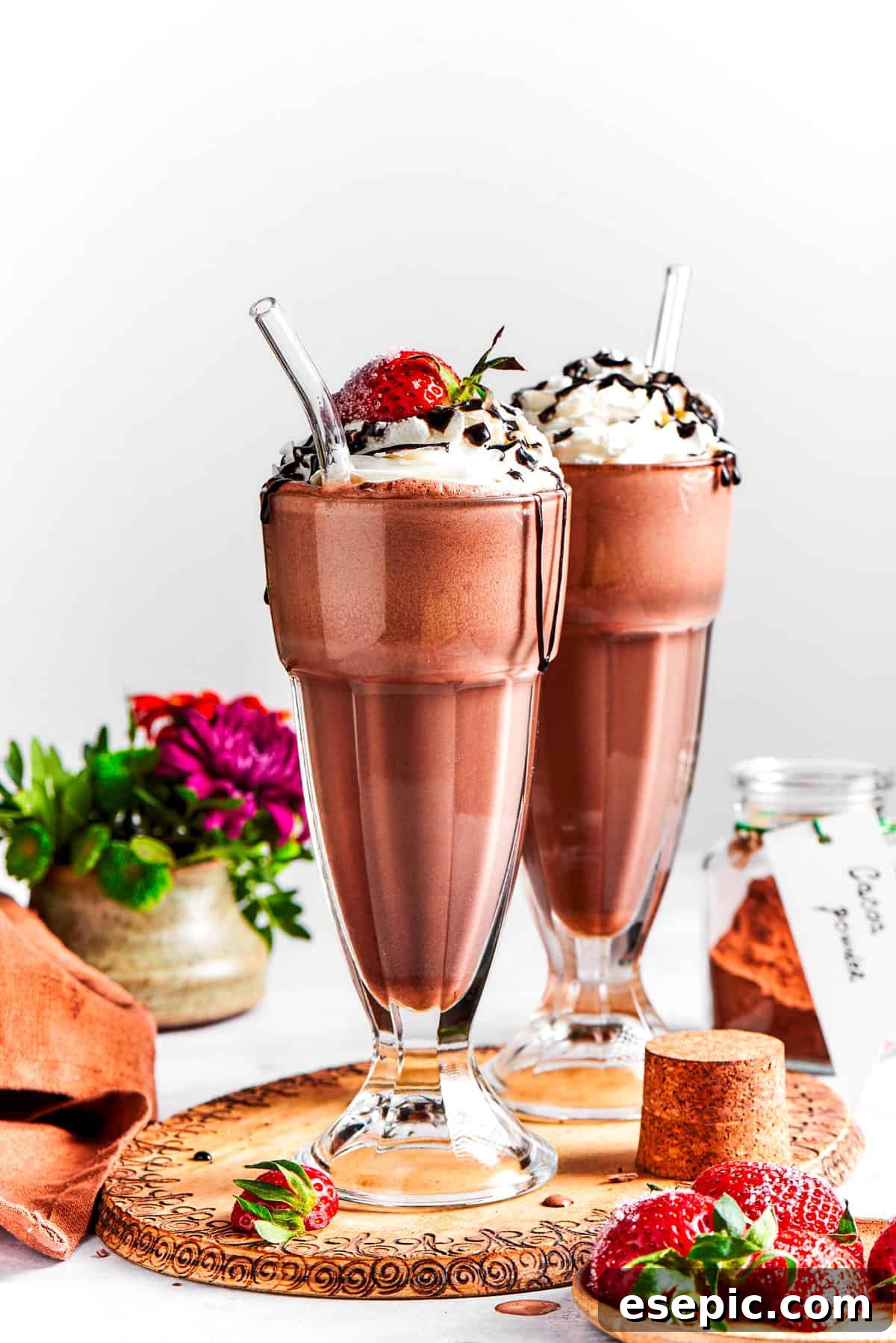 Two generous servings of chocolate milkshake, beautifully garnished with whipped cream, fresh strawberry slices, and a rich chocolate drizzle, resting on a rustic wooden tray.