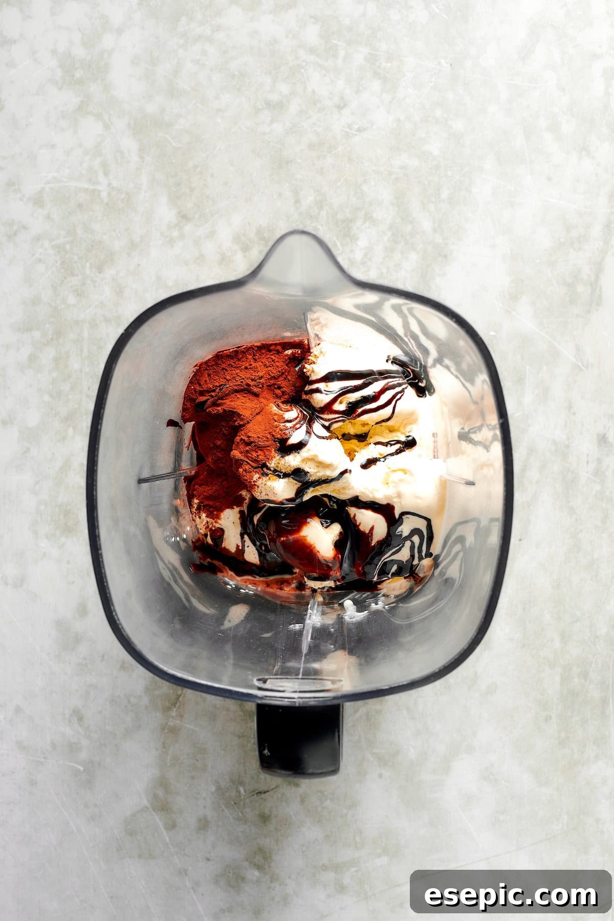 Vanilla ice cream, cocoa powder, chocolate syrup, and heavy cream are carefully added into a blender jar, preparing for blending.