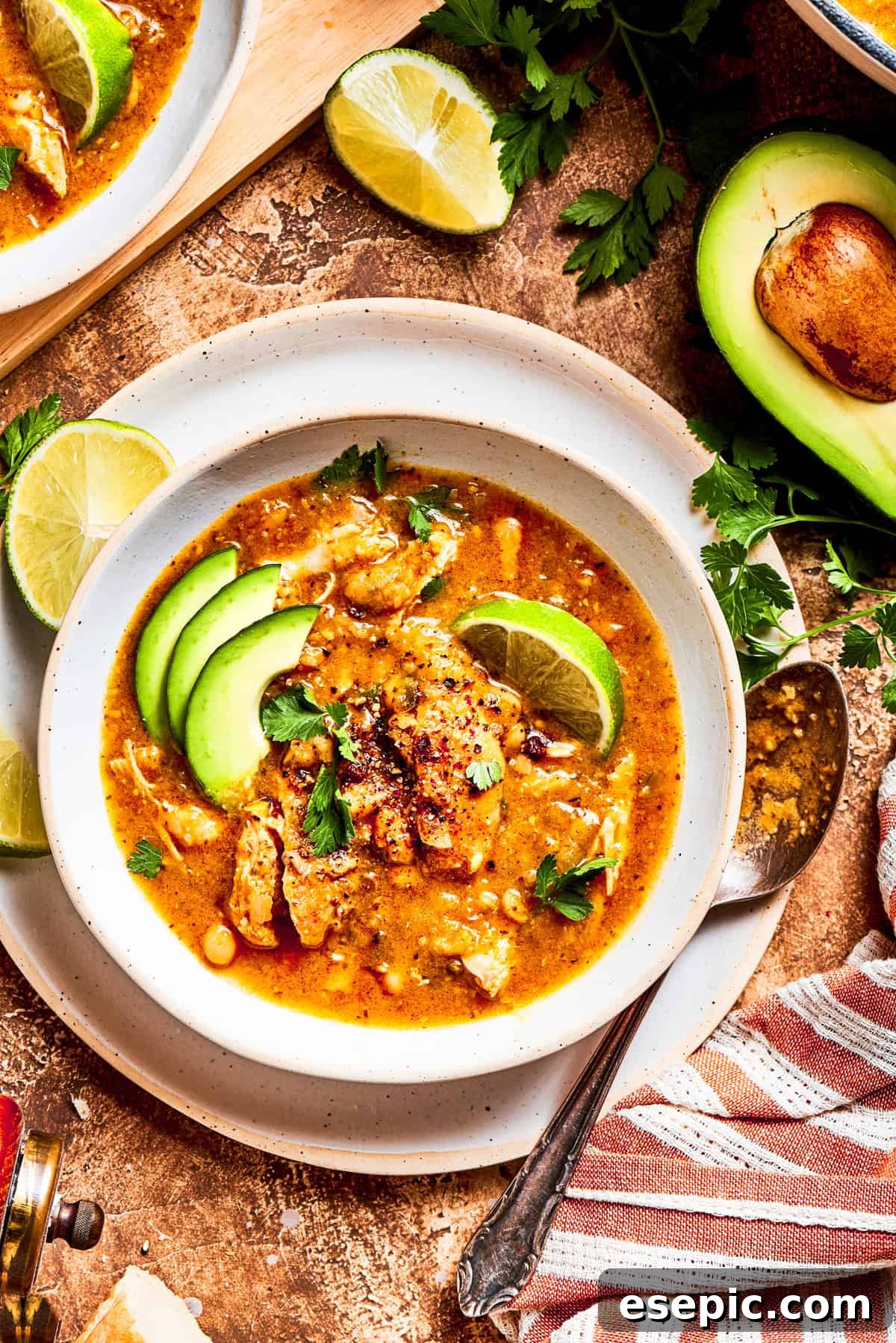 A bowl of vibrant spicy chicken soup, beautifully garnished with fresh avocado slices and bright lime wedges, ready to be enjoyed.