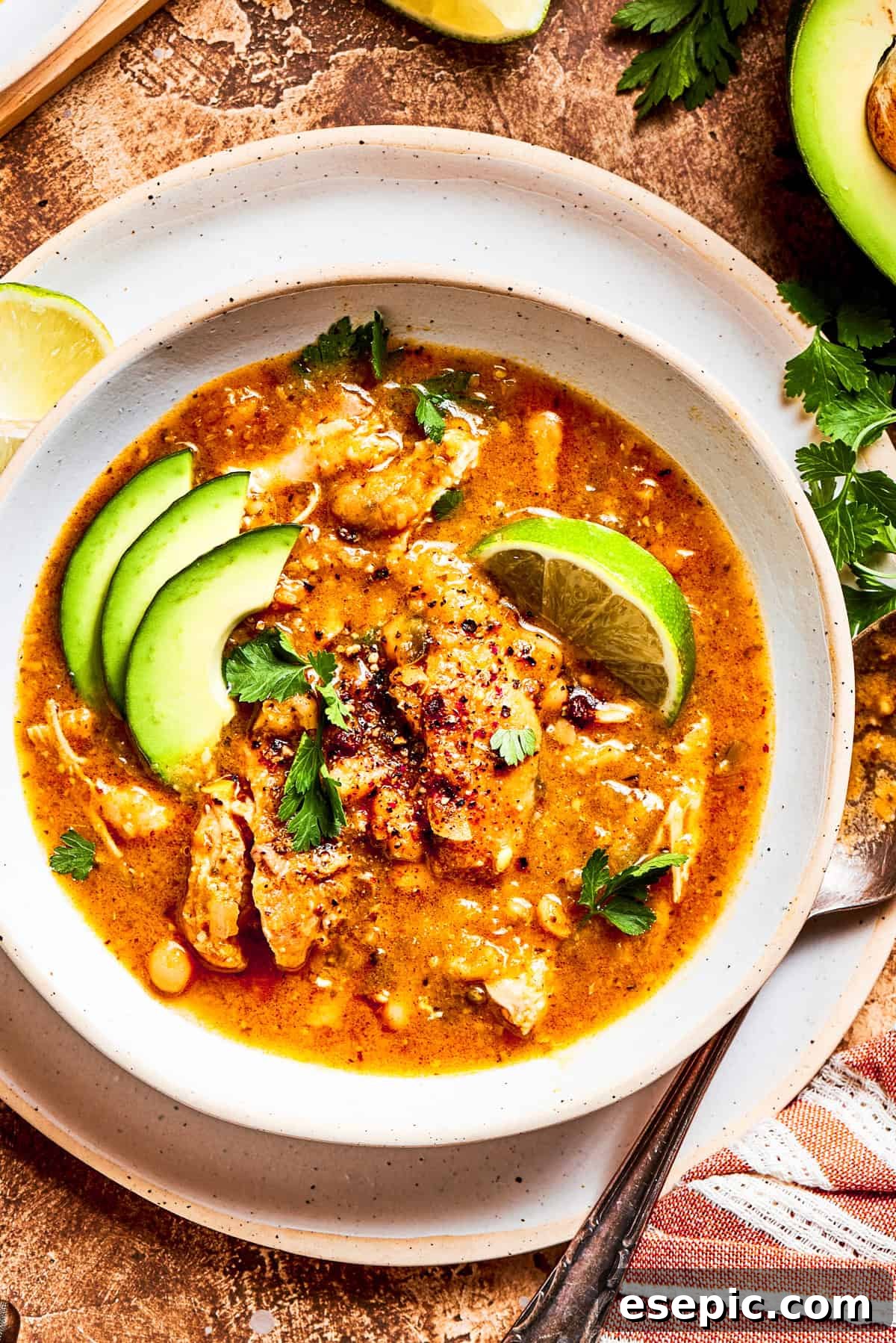 A vibrant bowl of spicy chicken soup, garnished with creamy avocado slices and bright lime wedges, ready for a delicious meal.