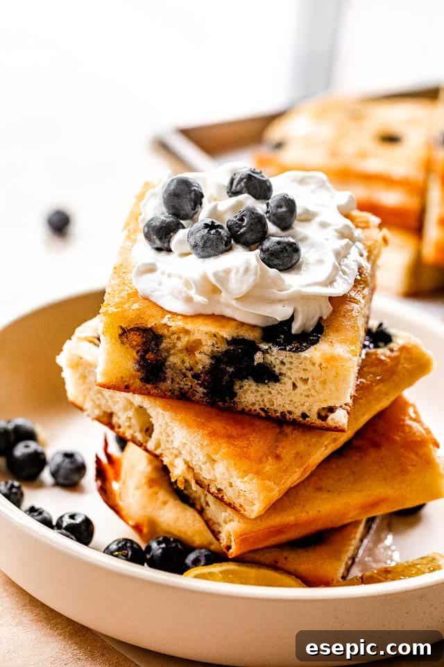 side shot of stacked sheet pan pancakes topped with whipped cream and blueberries
