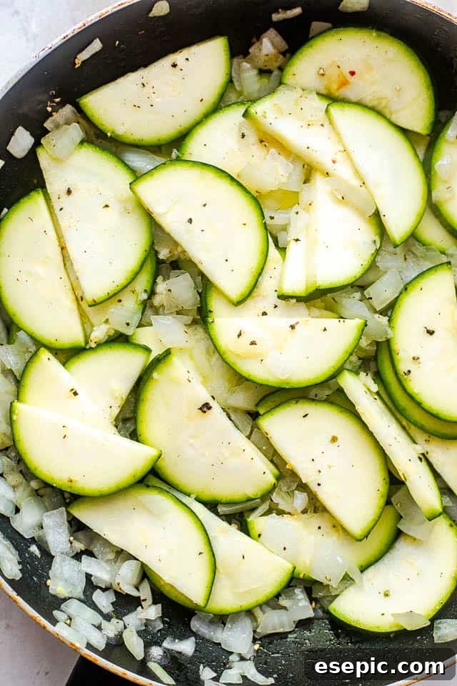 Velvety Zucchini Mushroom Pasta Bake 3 Close-up shot of zucchini slices and diced onion frying in a black skillet, creating the flavorful base for the pasta.