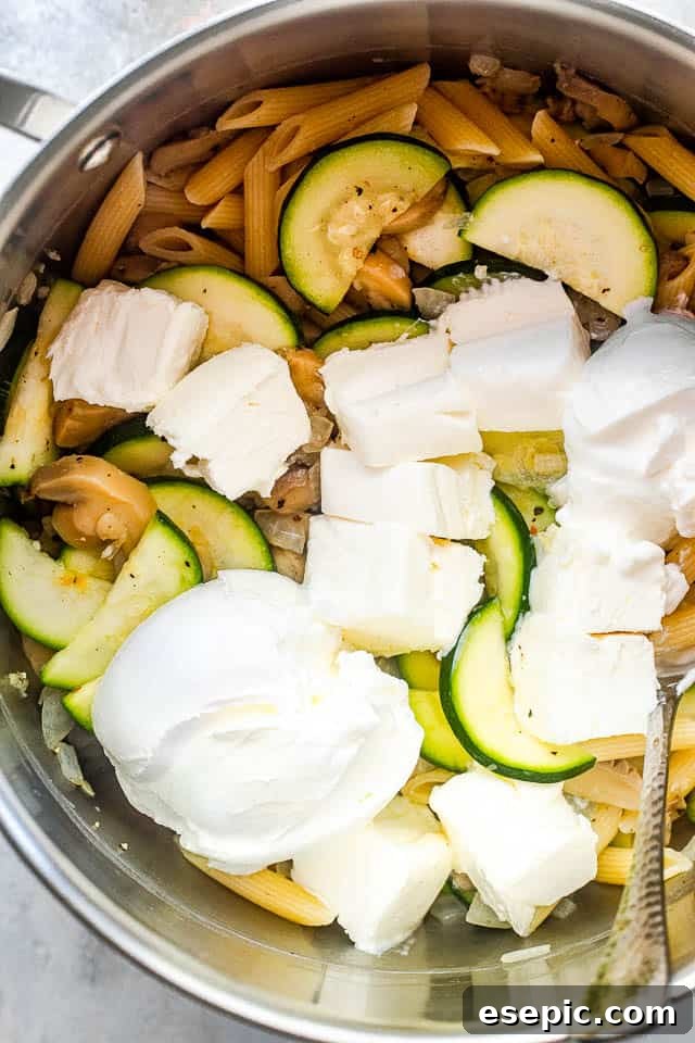 Velvety Zucchini Mushroom Pasta Bake 4 An overhead shot of a silver pot filled with cooked penne pasta, sautéed zucchini slices, and cubed cream cheese, ready to be mixed.