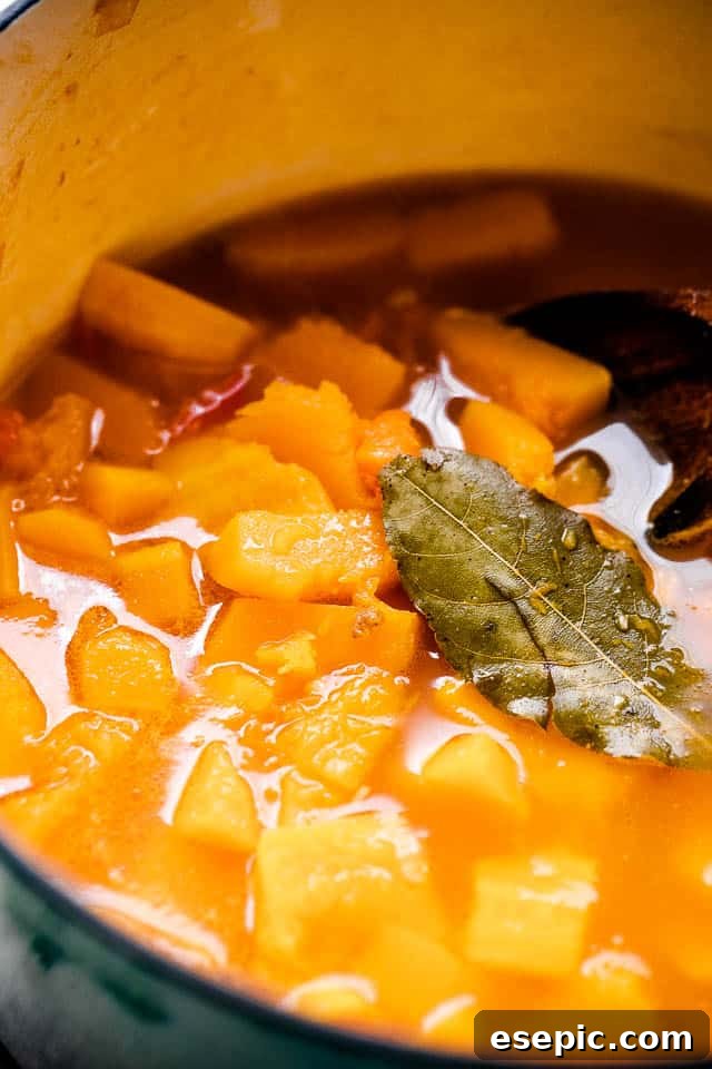 A rich and comforting soup prepared with spicy and bold Italian sausage, tender white beans, tangy diced tomatoes, and a base of pureed butternut squash. Diced Butternut squash simmering in broth, with a bay leaf set on top of the liquid in a Dutch oven.