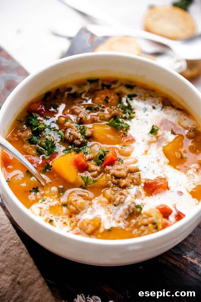 A rich and comforting soup prepared with spicy and bold Italian sausage, tender white beans, tangy diced tomatoes, and a base of pureed butternut squash. Side shot of butternut squash and sausage soup with a swirl of cream in a white soup bowl, ready to be enjoyed.