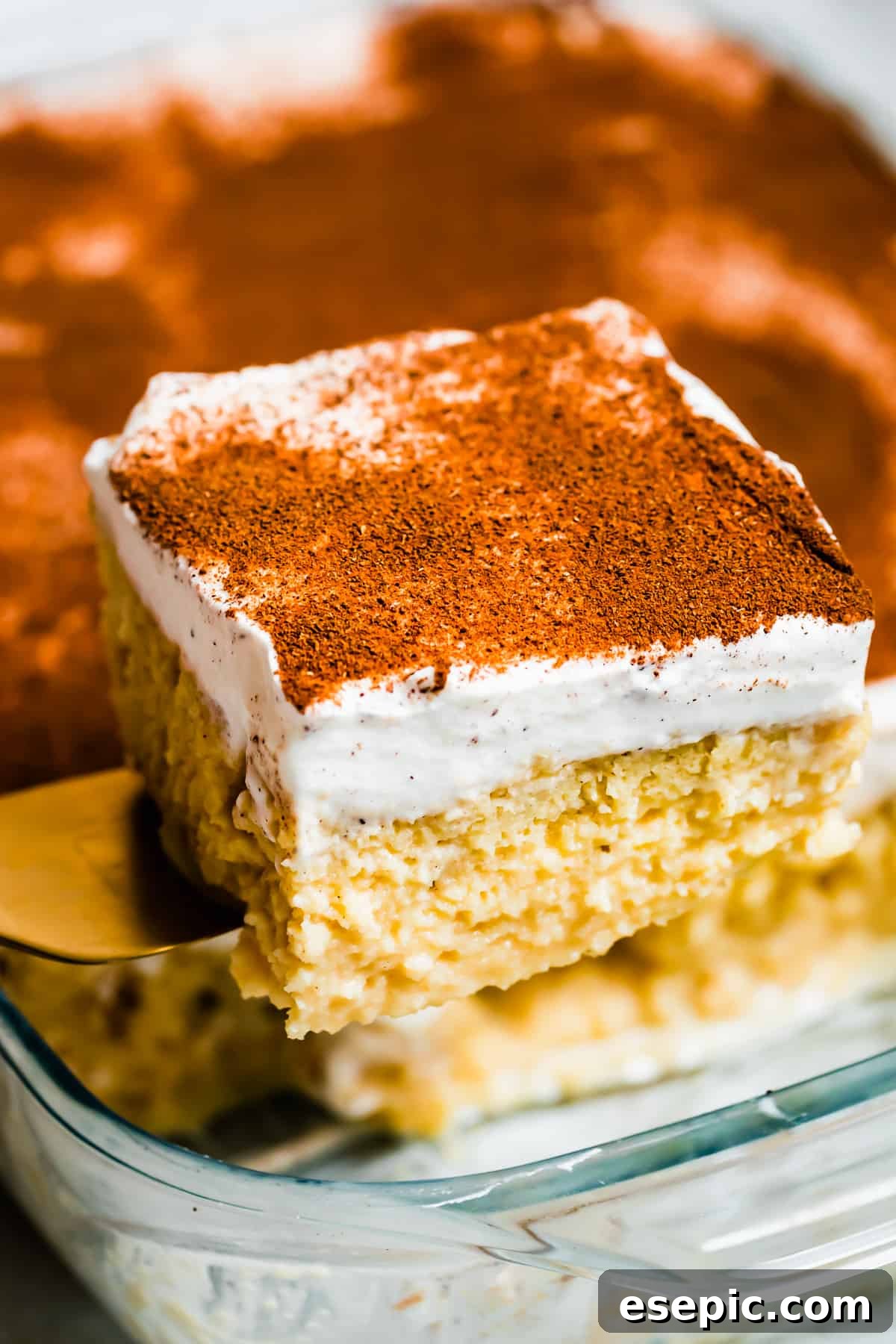 An easy Tres Leches Cake recipe with a sweet whipped cream and cinnamon topping. I use cake mix to make this Mexican dessert super simple! Lifting out a piece of tres leches cake from a glass baking dish, showcasing its moist texture and whipped cream topping.