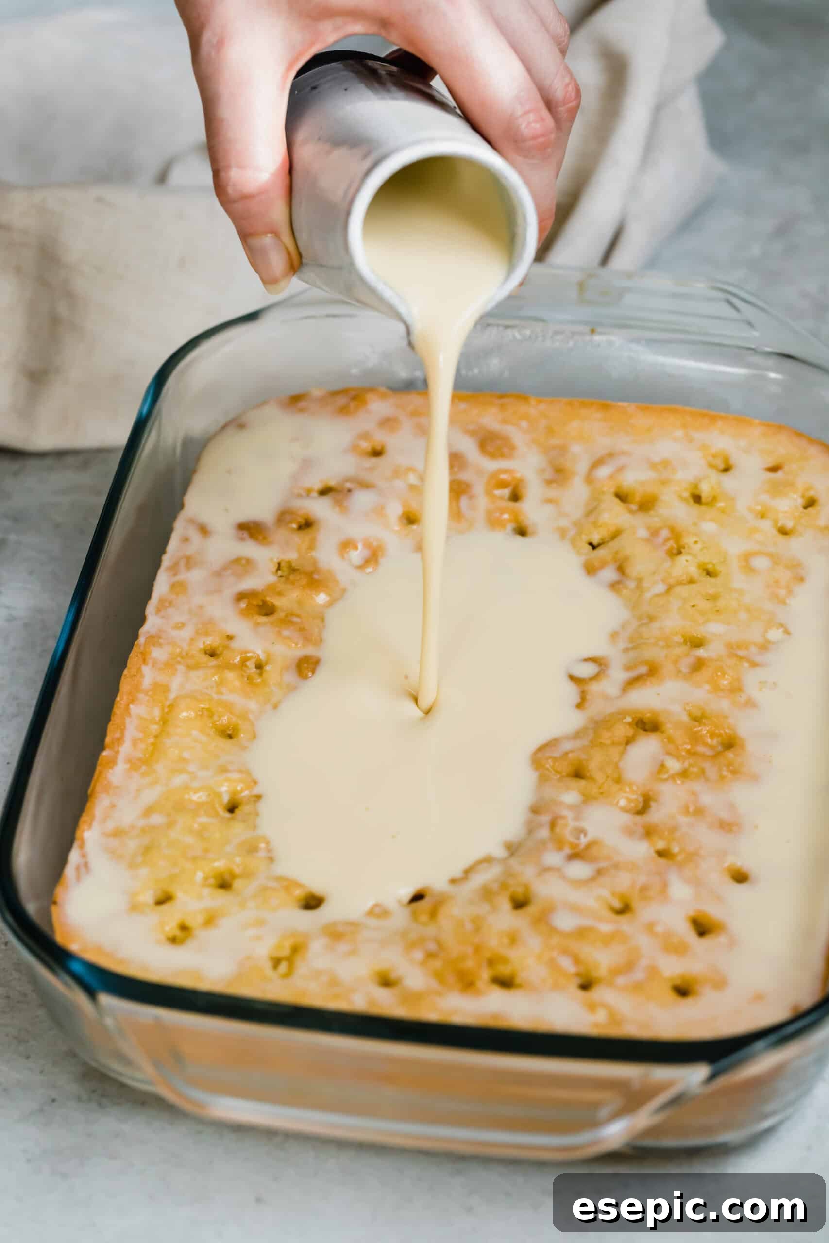 An easy Tres Leches Cake recipe with a sweet whipped cream and cinnamon topping. I use cake mix to make this Mexican dessert super simple! Close-up shot of the three milks mixture being poured over a baked sponge cake poked with holes, ready for soaking.