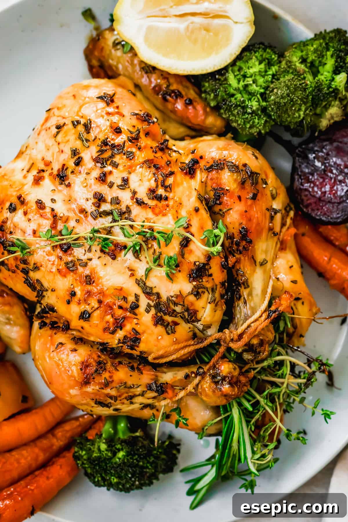 Overhead close-up shot of a perfectly roasted whole chicken served on a large white platter, surrounded by a colorful array of roasted root vegetables and broccoli florets, ready to be enjoyed.