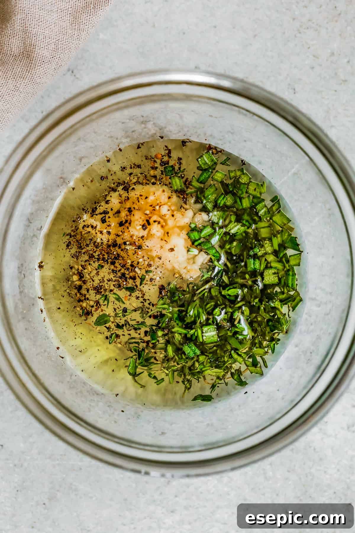Close-up shot of a small bowl containing olive oil, chopped fresh rosemary, thyme, minced garlic, salt, and pepper, ready to be mixed into a fragrant rub for the roast chicken.