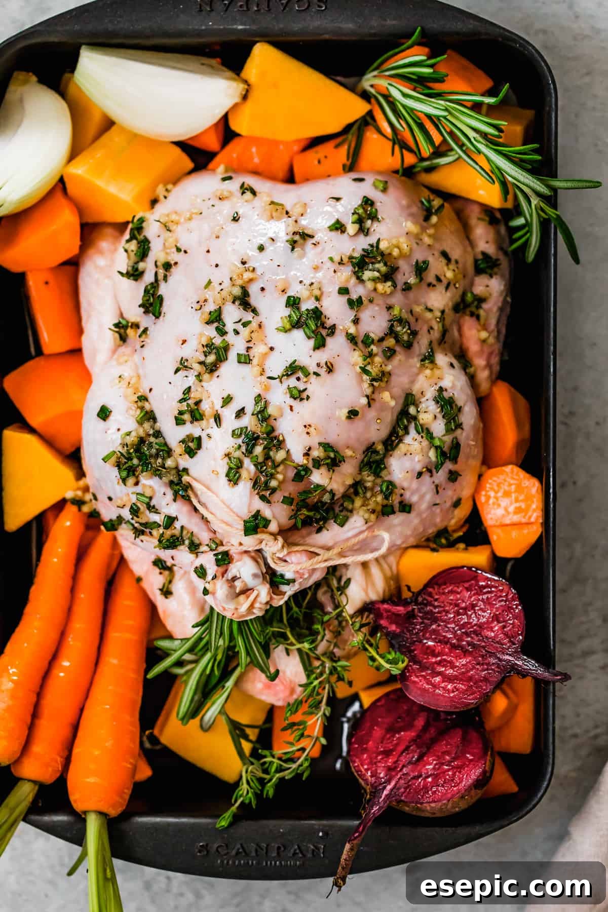 Raw whole broiler chicken neatly trussed in a baking pan, surrounded by a vibrant selection of raw root vegetables including butternut squash, sweet potatoes, carrots, and beets, ready for roasting.