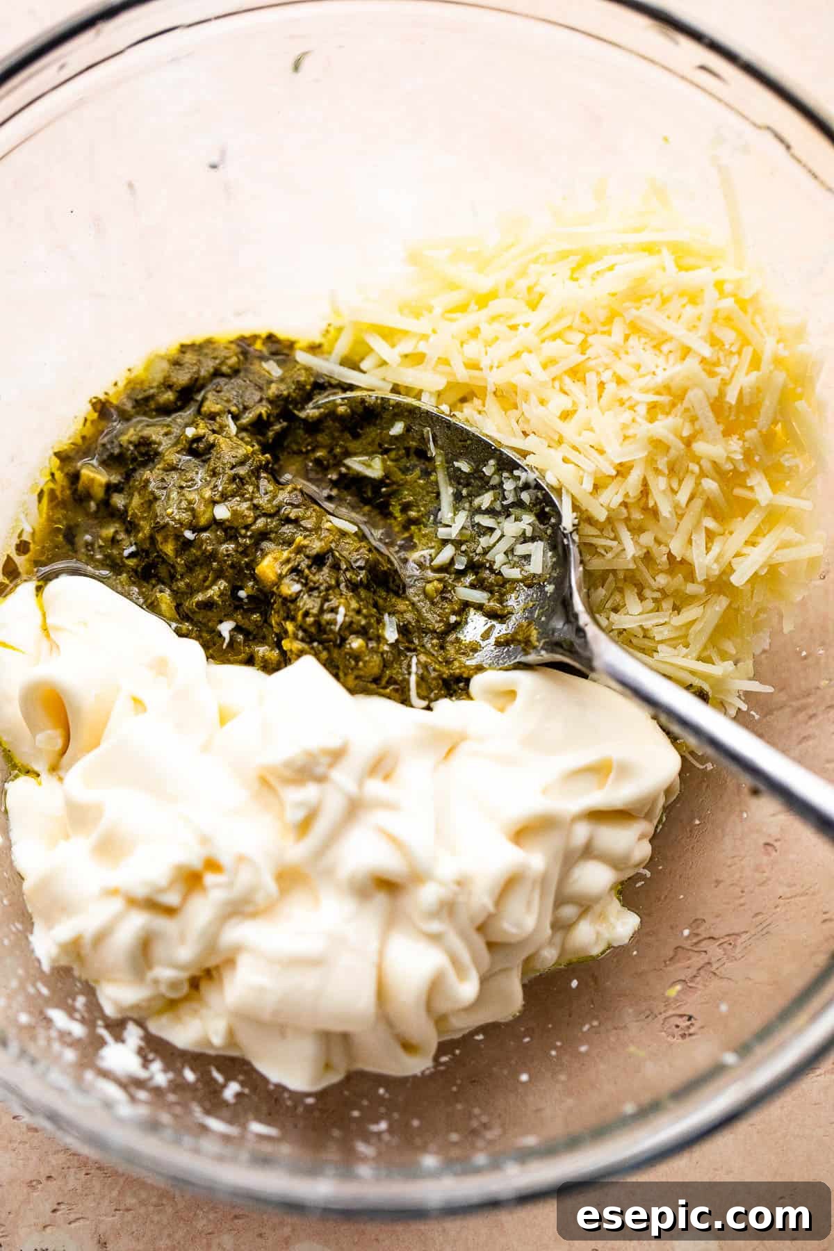 A bowl showing basil pesto, mayonnaise, and shredded parmesan cheese being mixed together to create the creamy sauce for the Caprese Chicken Casserole.