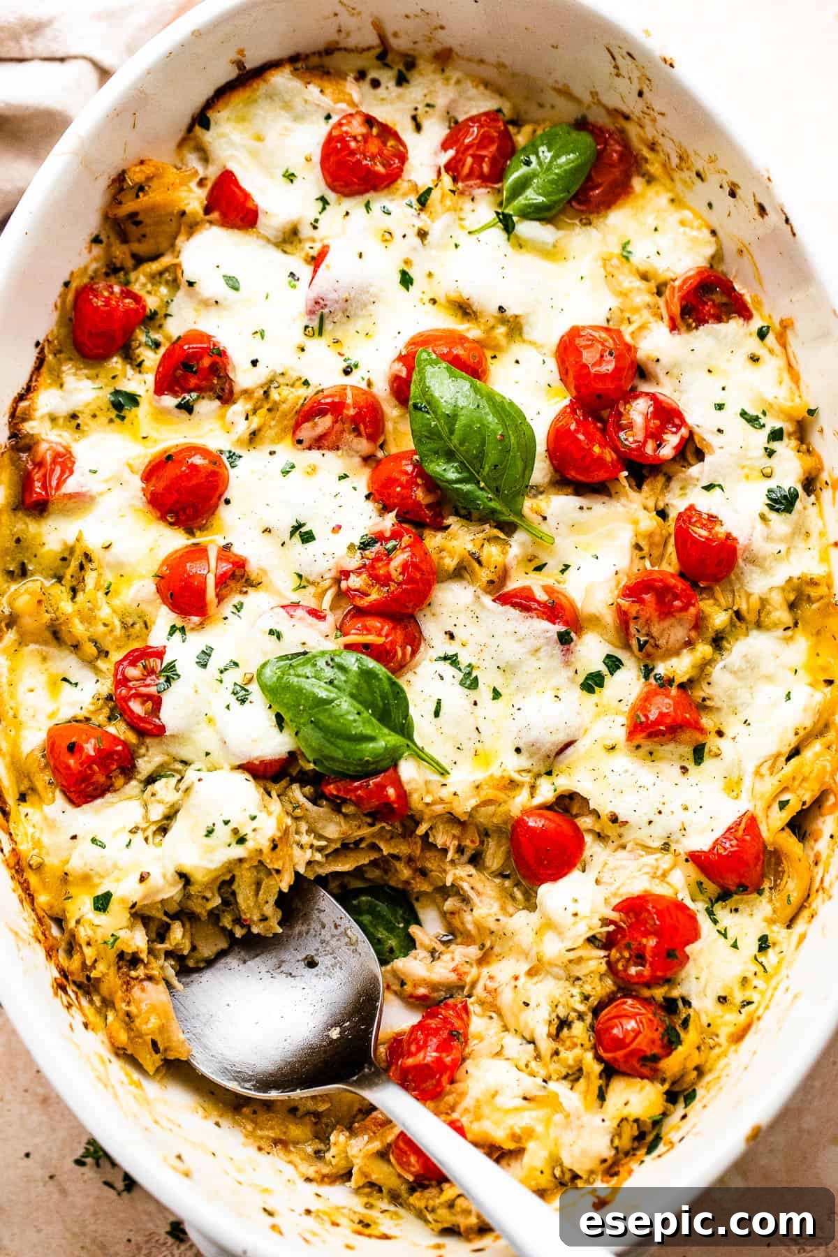 A spoon scooping out a serving of golden-brown Caprese Chicken Casserole from the white baking dish, showing the cheesy, saucy interior.
