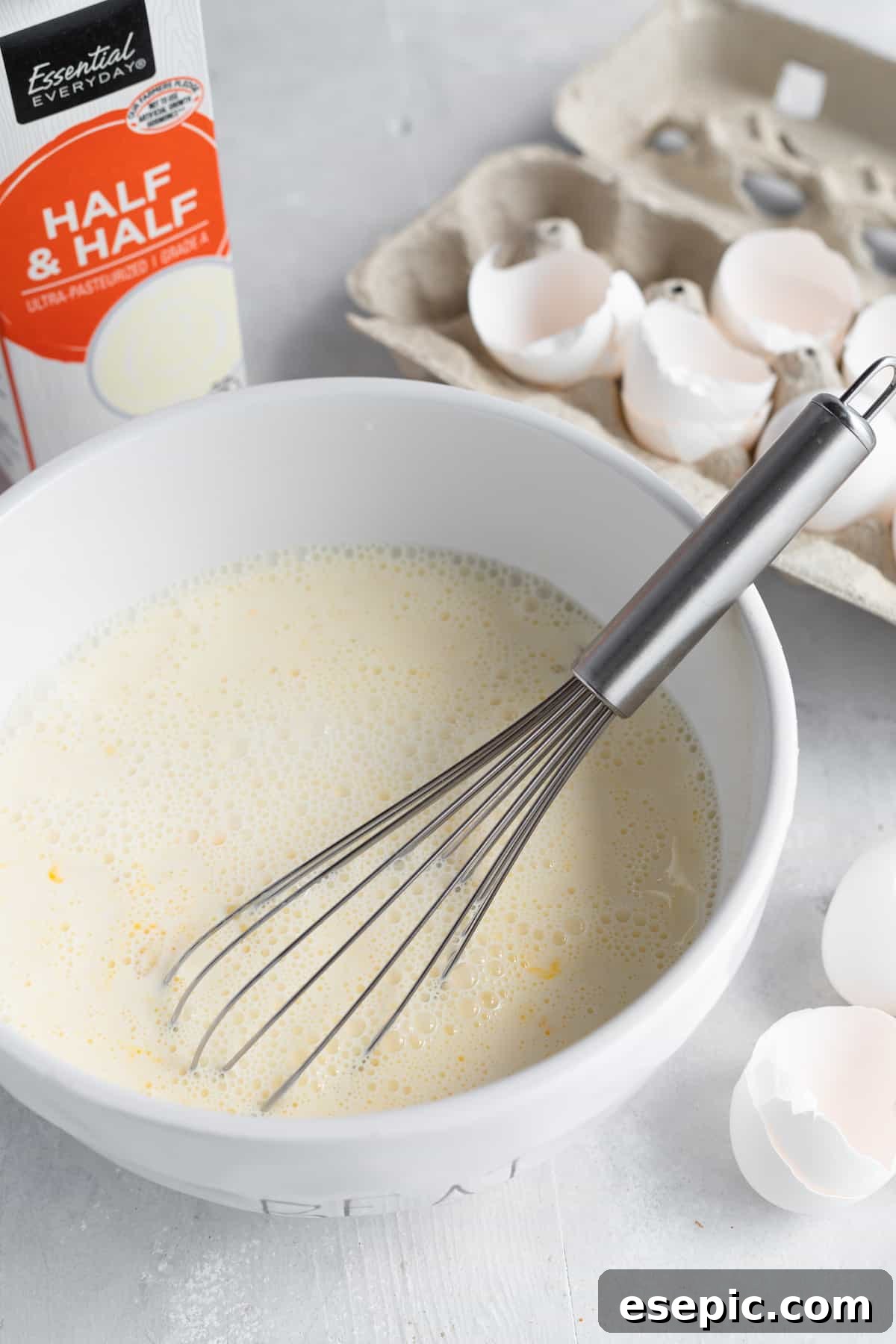 The Egg Mixture in a White Bowl with a Metal Whisk and Egg Shells in the Background