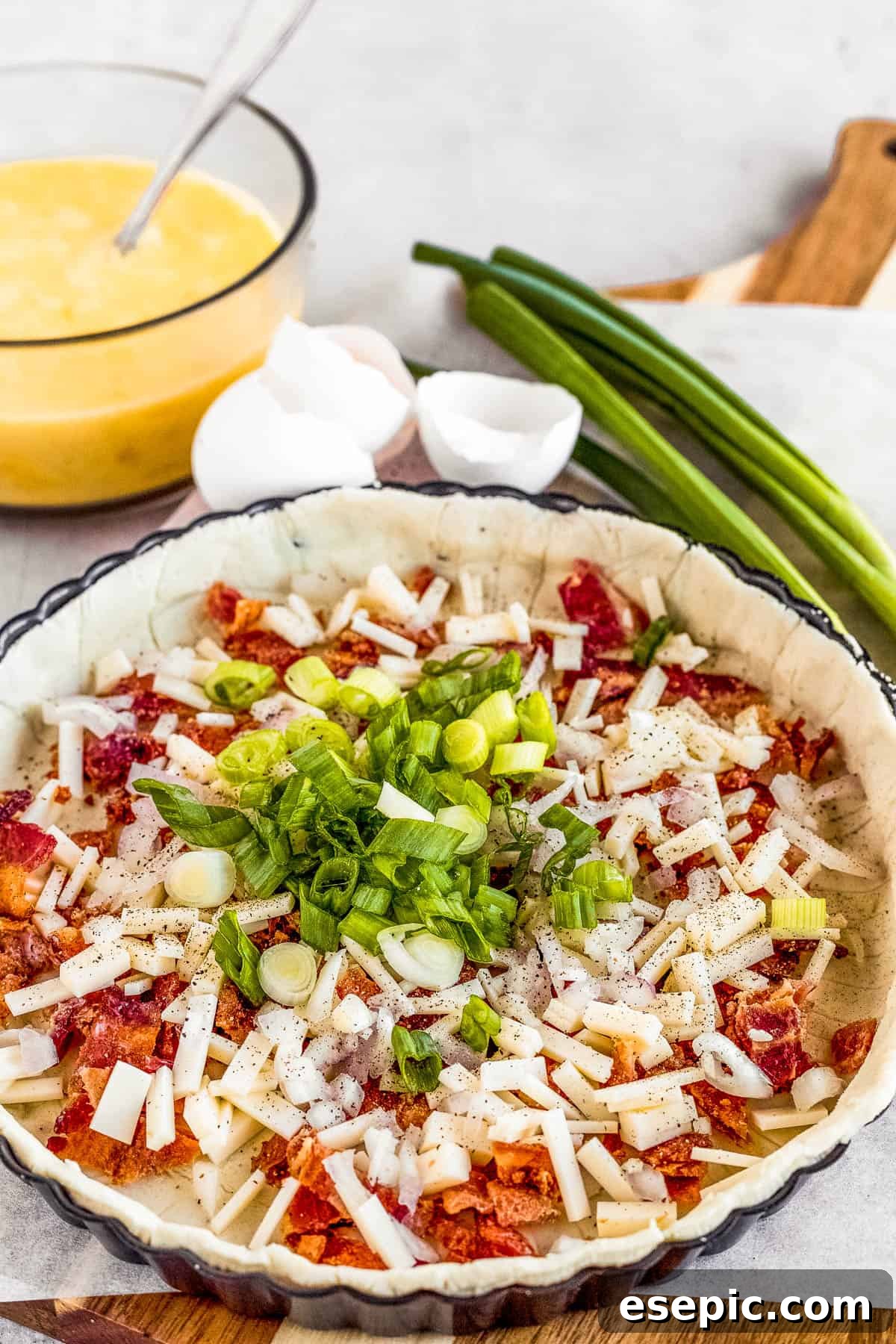 Effortlessly Delicious Bacon Quiche Lorraine 5 Chopped onions, shallots, and bacon bits carefully arranged inside a pie crust, with the whisked egg mixture visible in the background, ready to be poured.