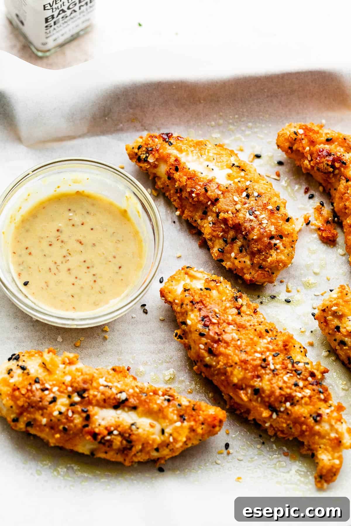 Perfectly baked Everything Bagel Chicken Tenders showcased on a pristine white background, accompanied by a small bowl of savory dipping sauce.