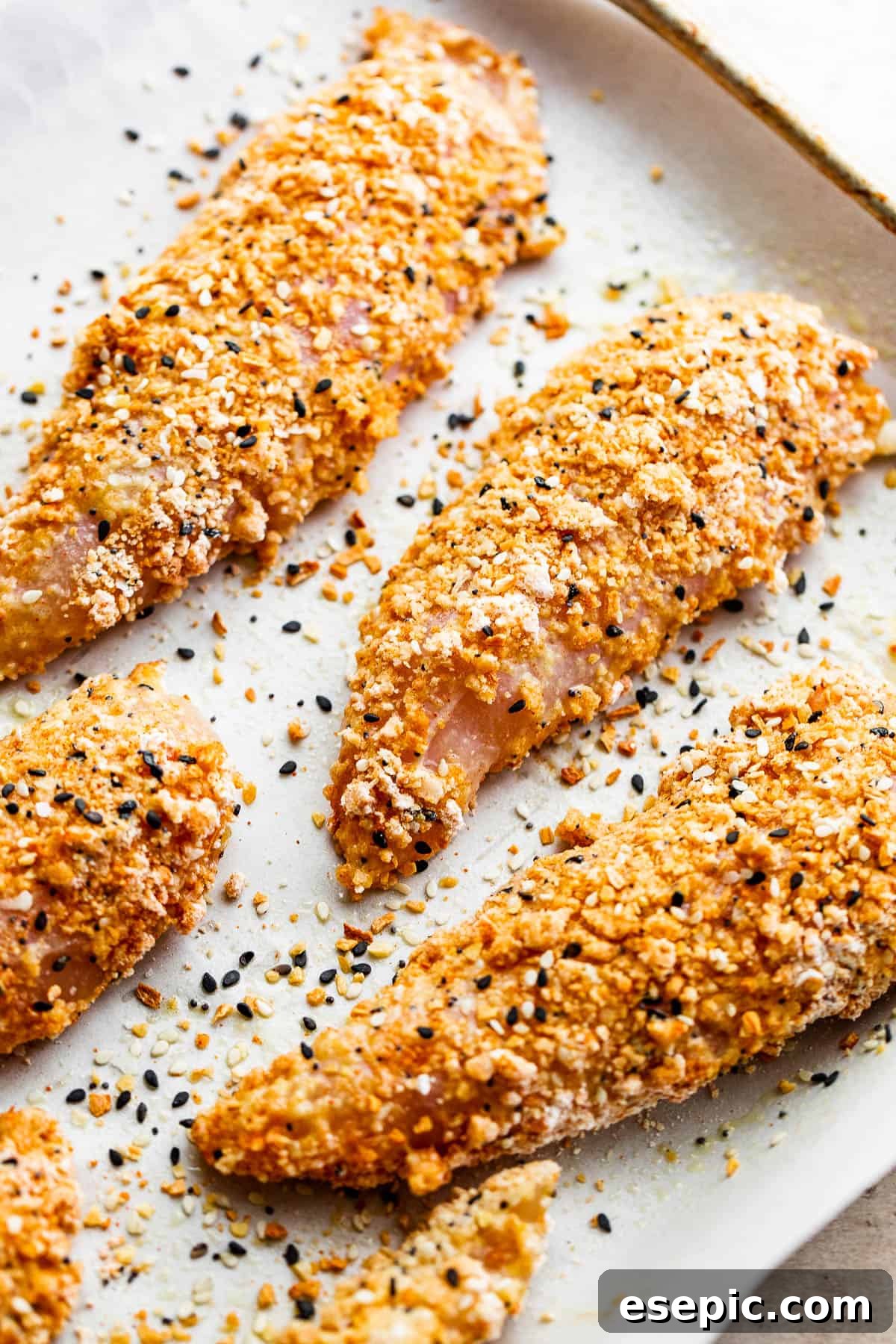 Raw chicken tenders generously covered with a vibrant and aromatic everything bagel seasoning mix, ready for baking.