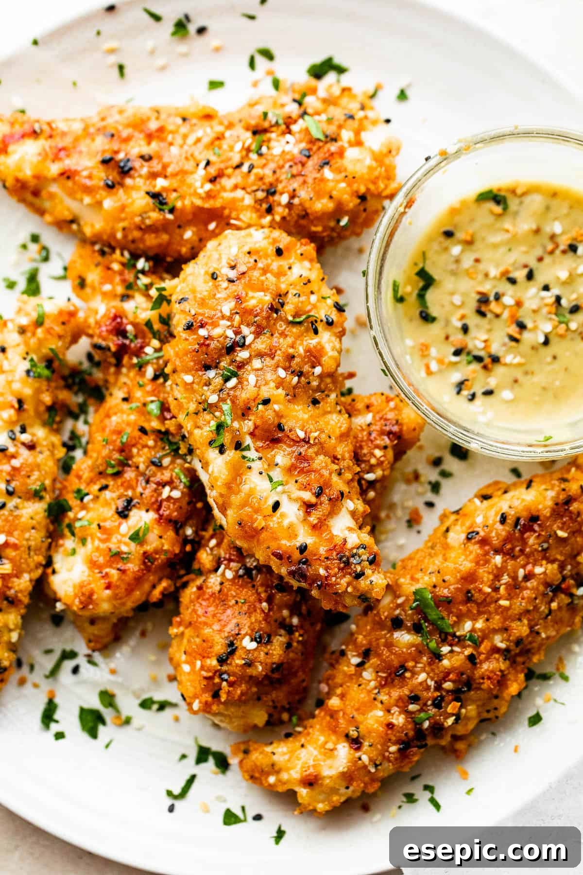 A close-up shot of six beautifully arranged Everything Bagel Chicken Tenders on a round plate, served with a side of honey mustard dipping sauce.