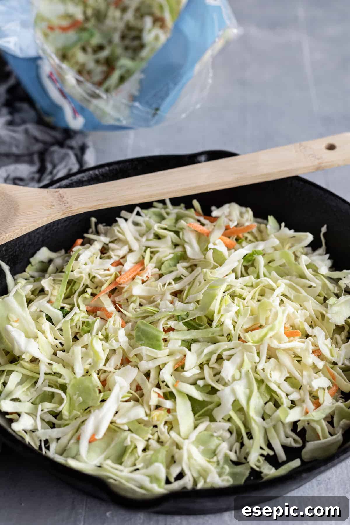 Cabbage coleslaw mixture gently wilting in a frying pan with a wooden spoon.