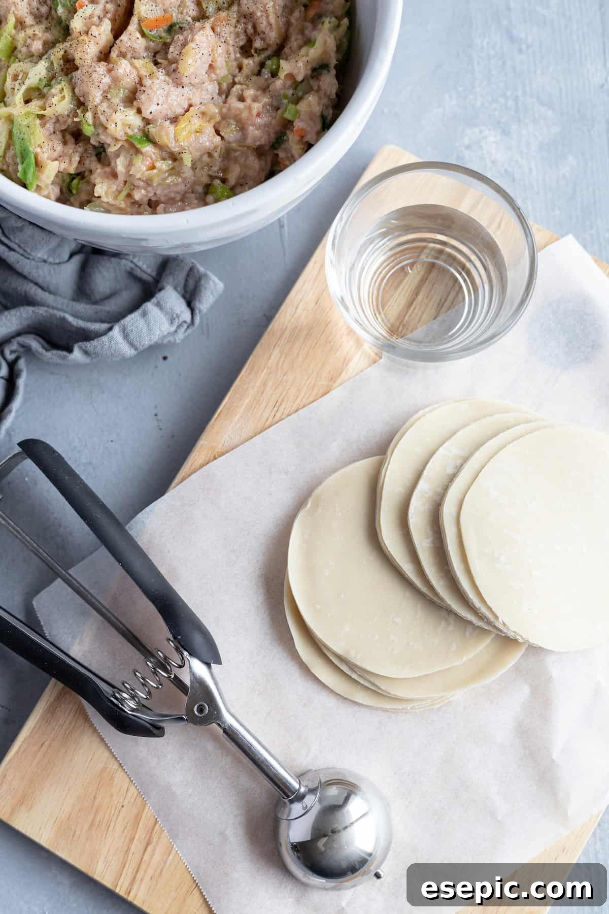 A tidy workspace for making potstickers, featuring a pile of round dumpling wrappers, a bowl of filling, and a small cup of water.