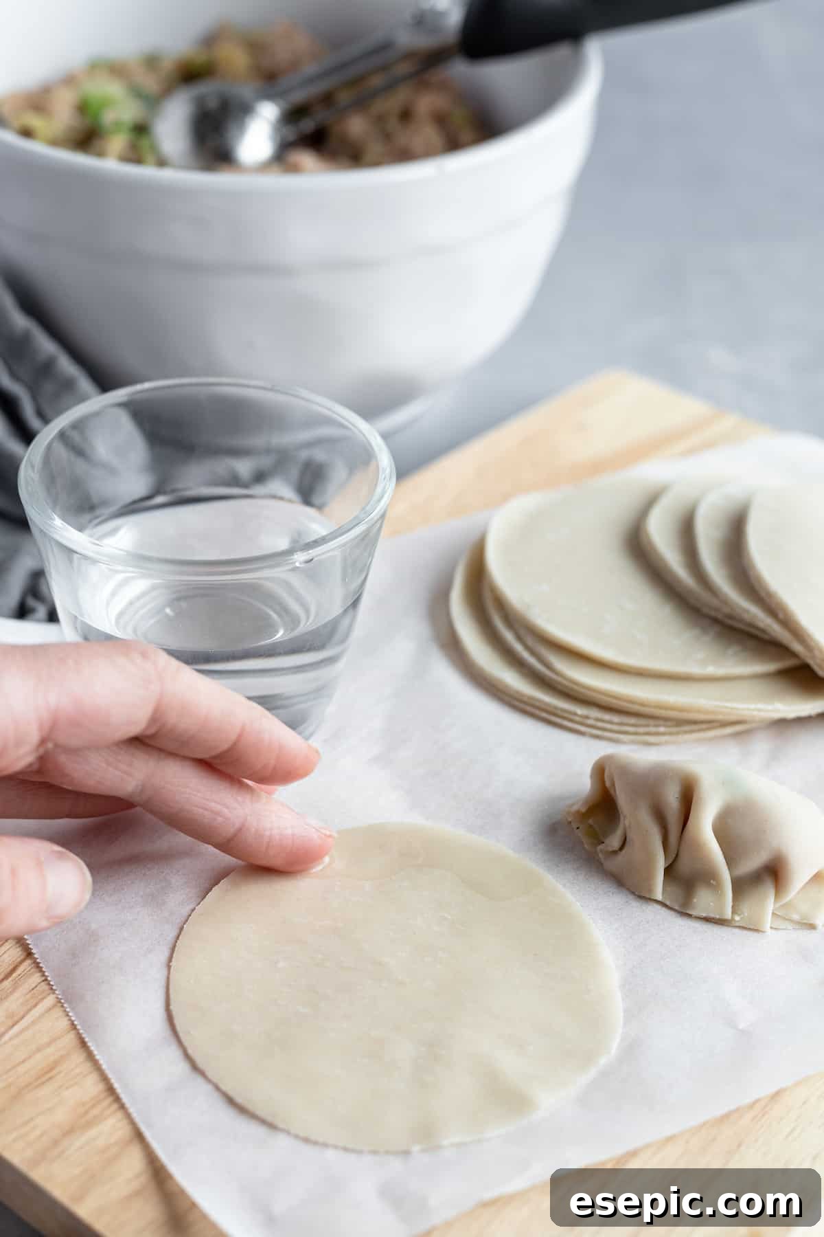 A finger gently moistening the edge of a round Asian dumpling wrapper on a flat surface.