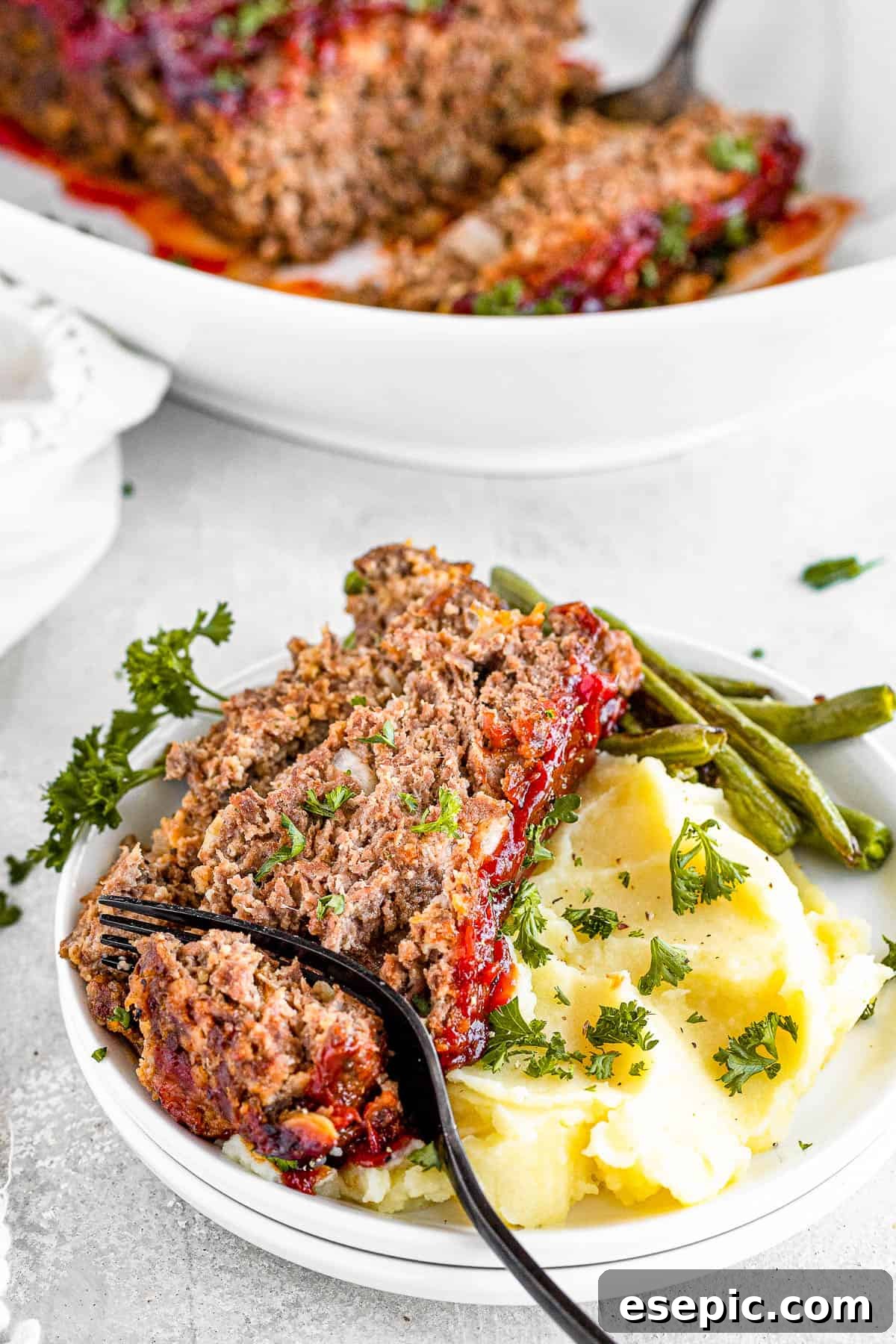 A family-friendly classic, this easy meatloaf recipe gets extra flavor from barbecue sauce, plenty of garlic, and Italian seasoning. A Fork Digging Into a Slice of Meatloaf on a Plate with Potatoes and Veggies