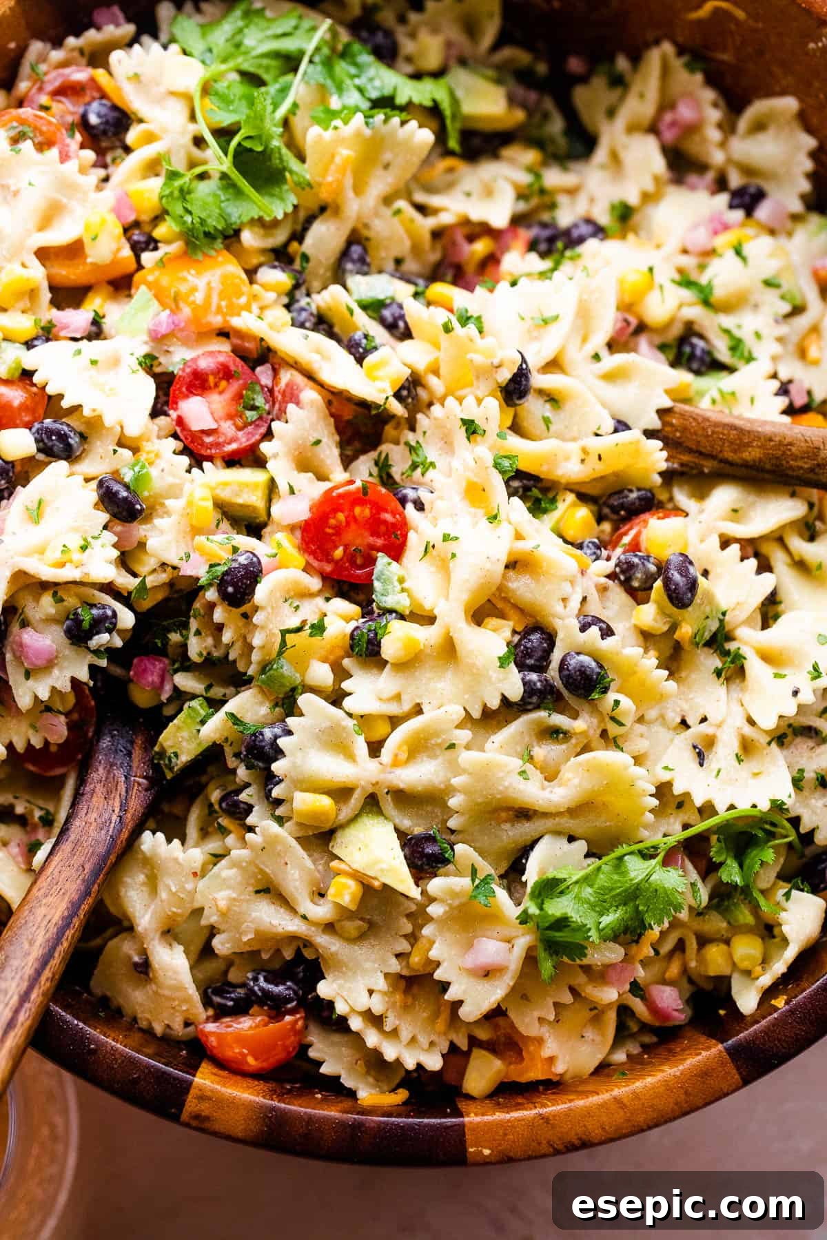 A close-up view of the vibrant Fiesta Pasta Salad, showcasing the diverse colors and textures of pasta, black beans, corn, avocado, and tomatoes, served in a wooden bowl with wooden spoons.
