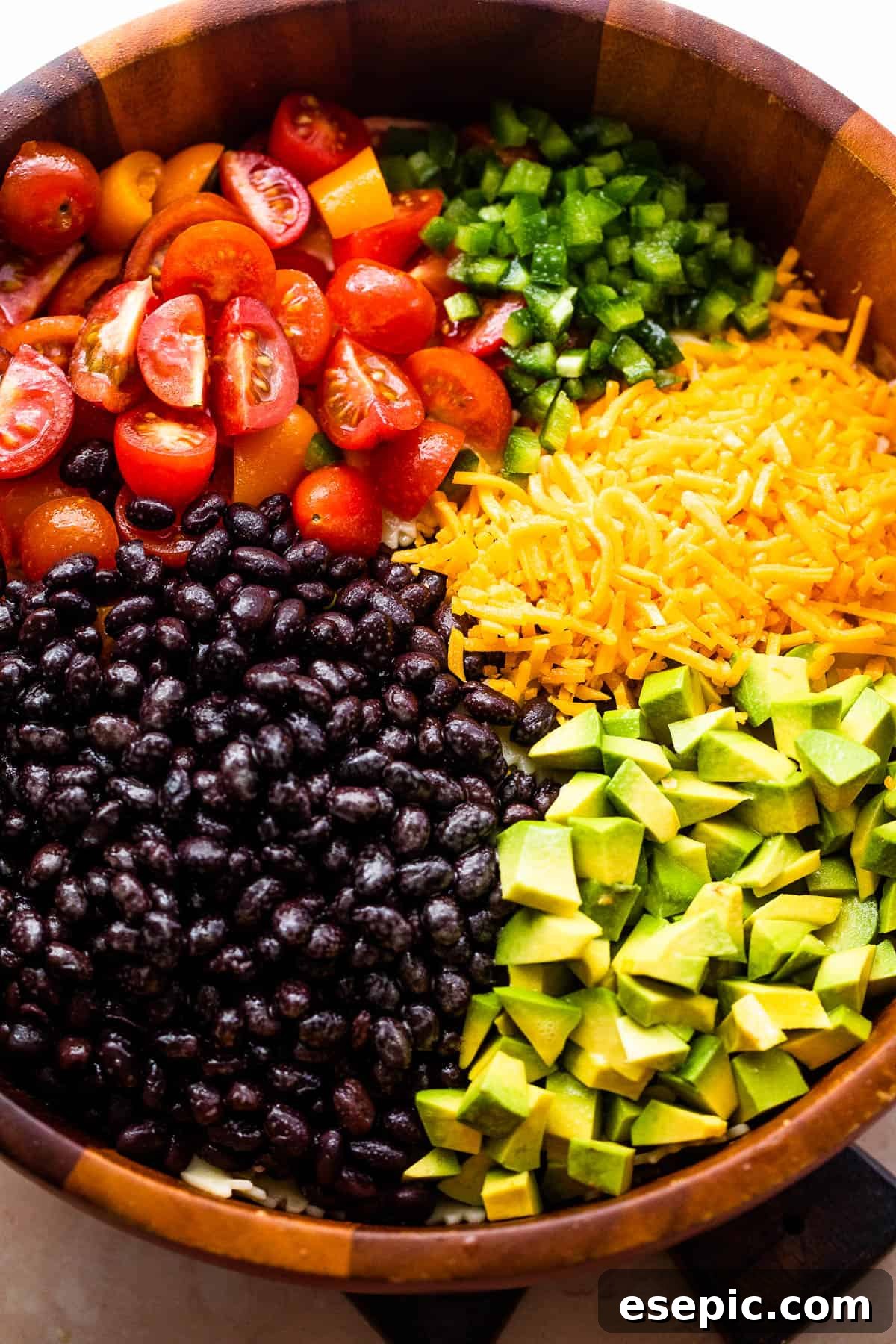 A wooden bowl filled with an assortment of vibrant Fiesta Pasta Salad ingredients including diced tomatoes, creamy avocado, shredded cheddar cheese, and wholesome black beans.