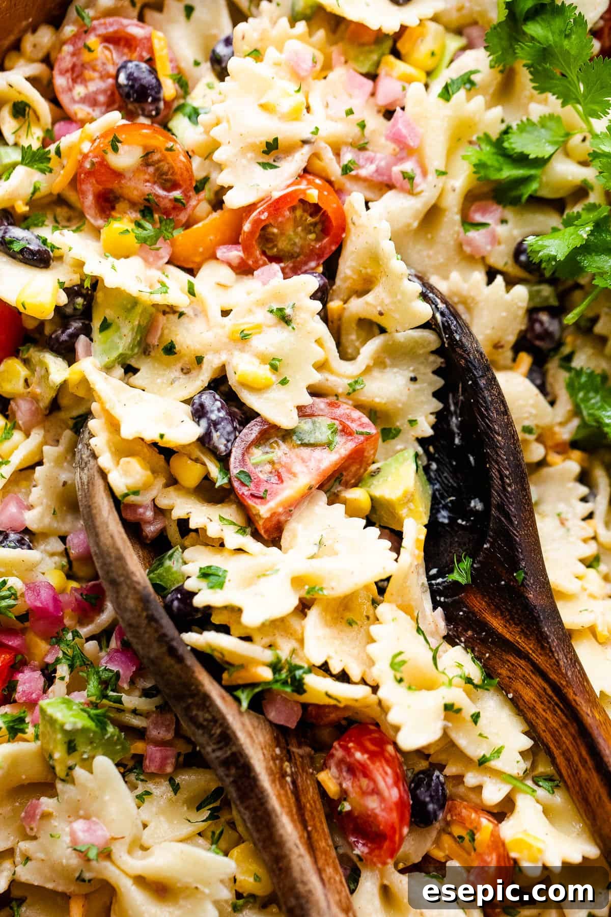 A close-up of the Fiesta Pasta Salad, rich with avocado, black beans, corn, and pasta, in a wooden bowl with serving spoons, inviting a taste.