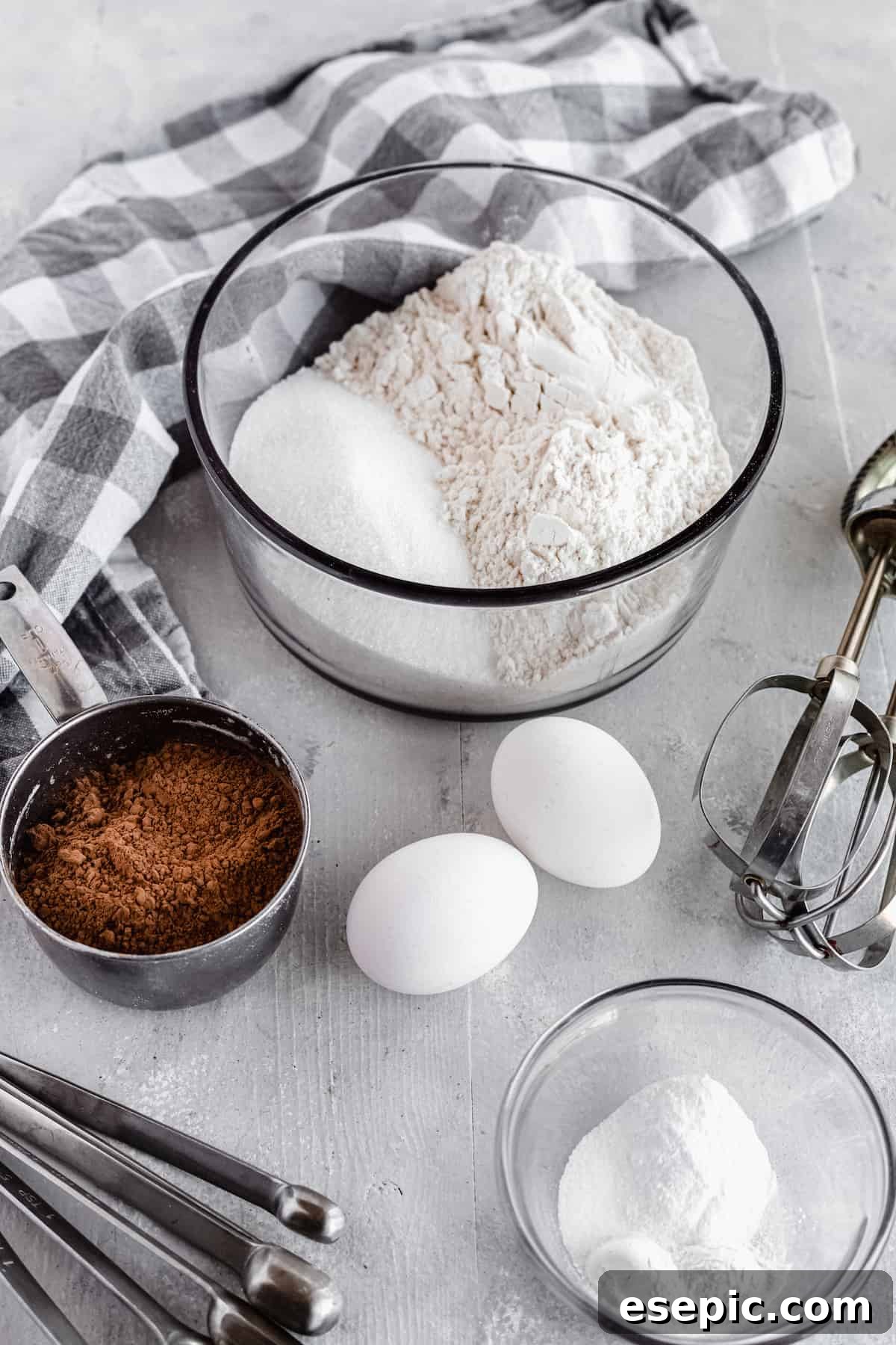 Key baking ingredients for chocolate cake, including cocoa powder, eggs, baking soda, baking powder, flour, and sugar, neatly arranged on a gray countertop.