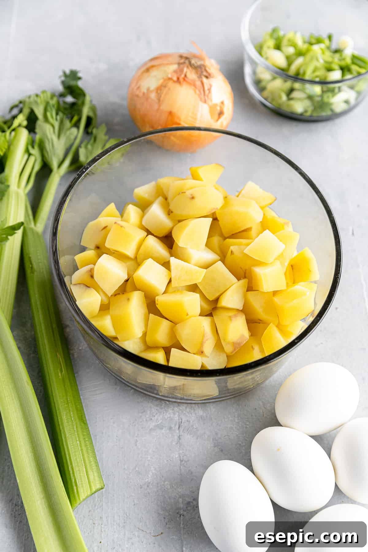Organized Ingredients for Classic Potato Salad, Featuring Chopped Yukon Gold Potatoes, Hard-Boiled Eggs, and a Yellow Onion on a Gray Countertop