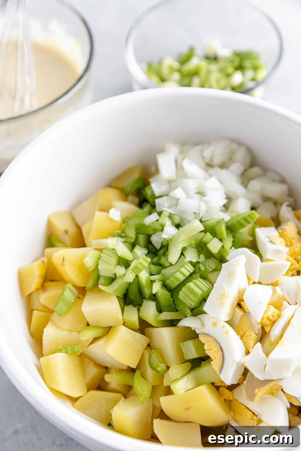 A White Mixing Bowl Containing Uncombined Cooked Potatoes, Diced Celery, Onion, and Hard-Boiled Eggs