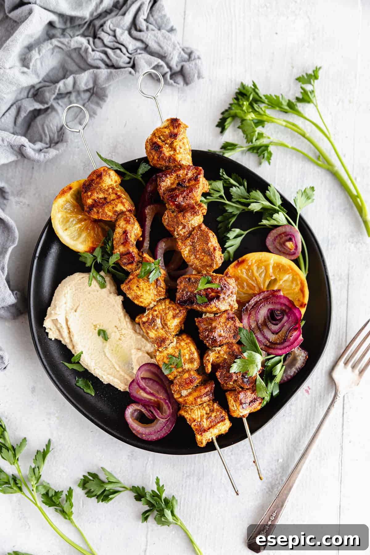 A Black Plate Containing Two Chicken Skewers Along with Various Fruits and Vegetables with a Fork Beside it
