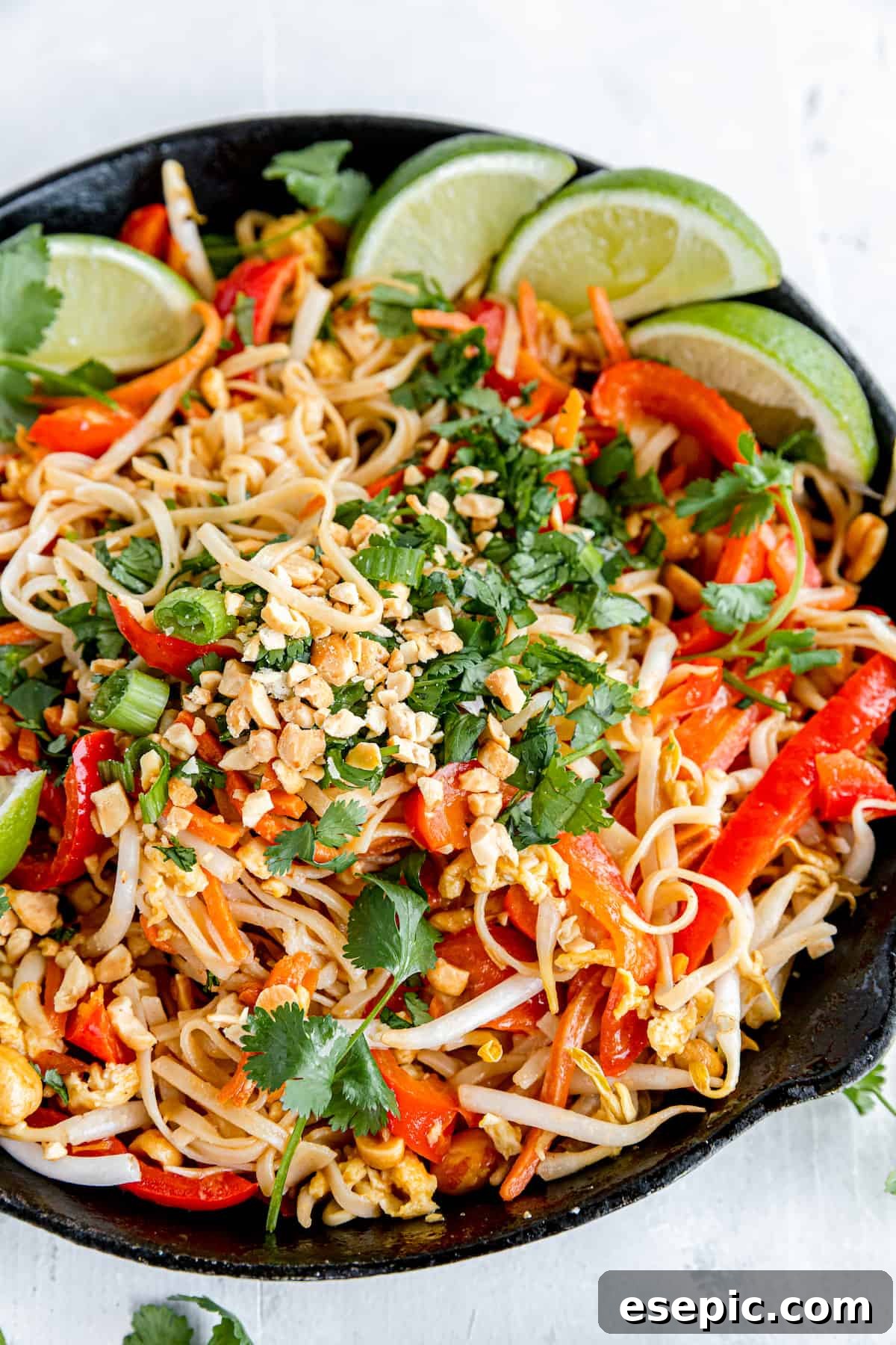 A Black Skillet Filled with Pad Thai on Top of a White Dinner Table, garnished with fresh lime, cilantro, and peanuts.