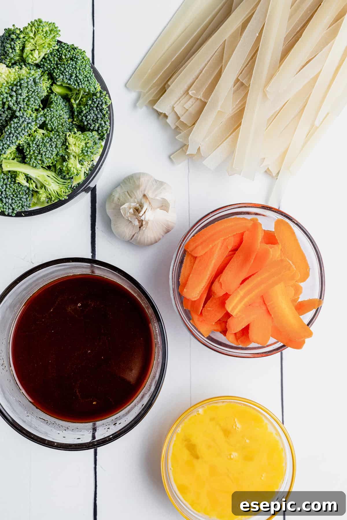 Various ingredients for Pad See Ew, including broccoli, flat rice noodles, and sliced carrots, neatly arranged on a white picnic table.