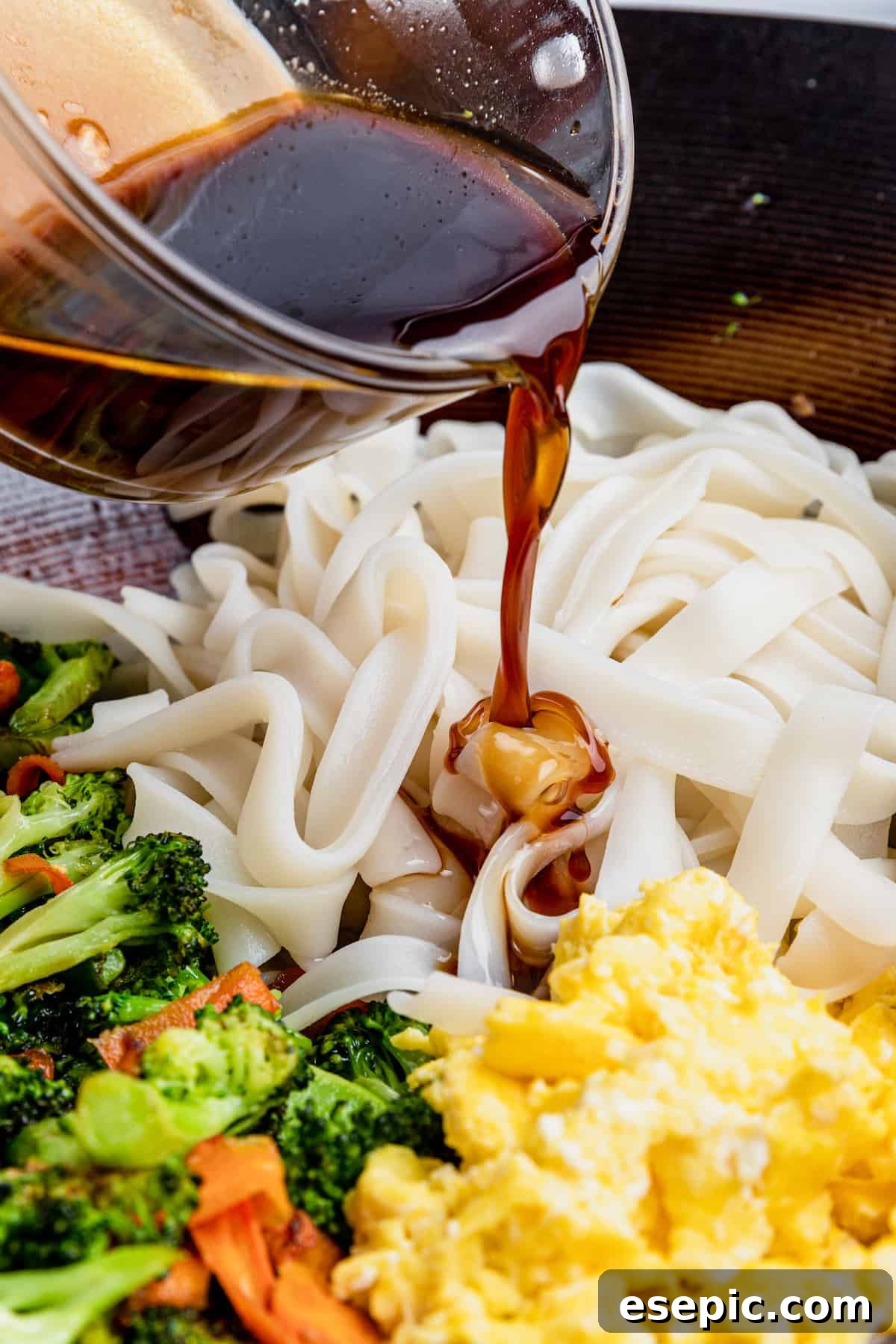 Rich Pad See Ew sauce being poured over the cooked noodles, eggs, and vegetables in a mixing bowl, prior to final stir-frying.