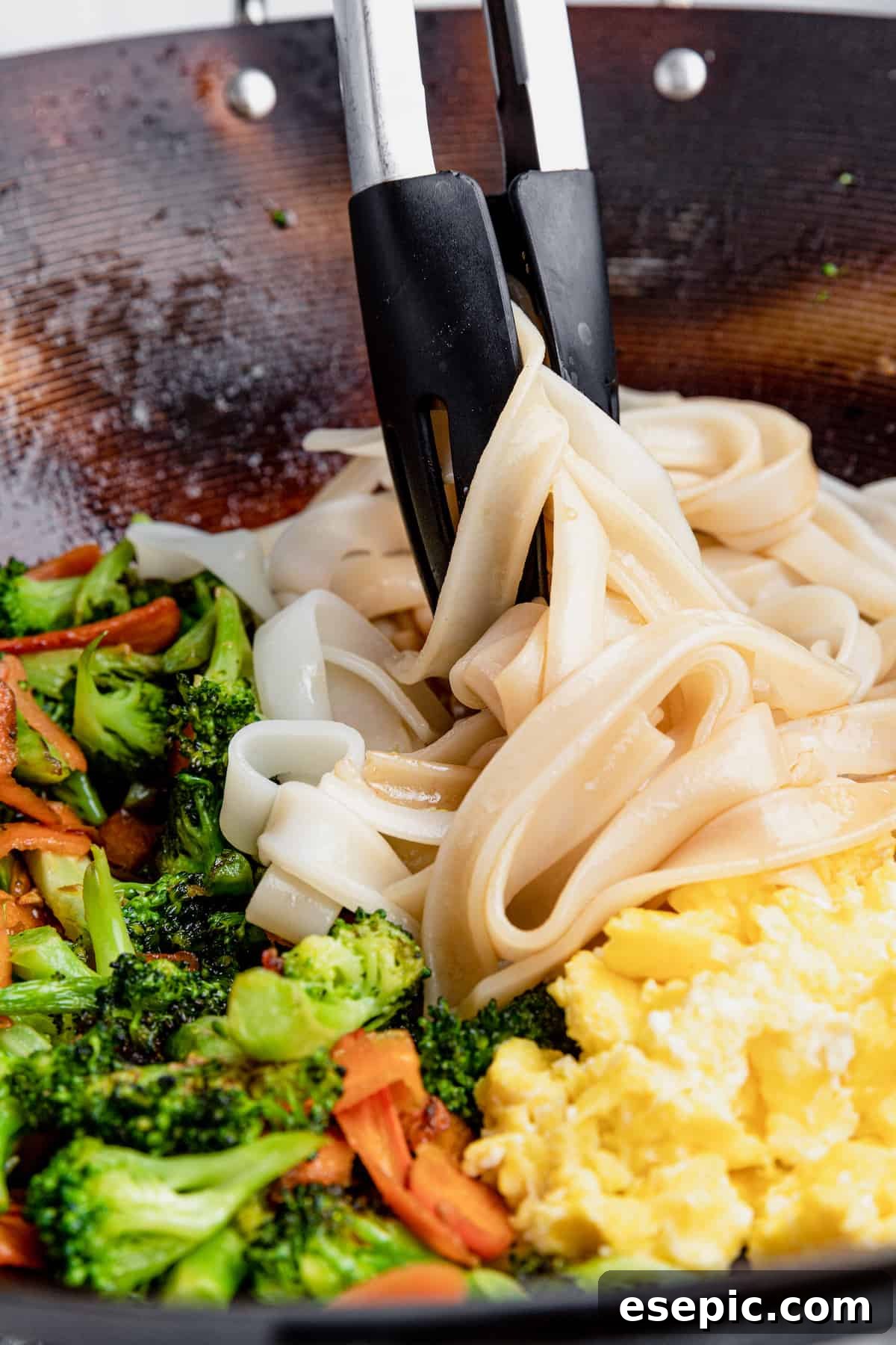 A pair of tongs gently mixing the noodles, eggs, and vegetables with the savory sauce, ensuring even coating.
