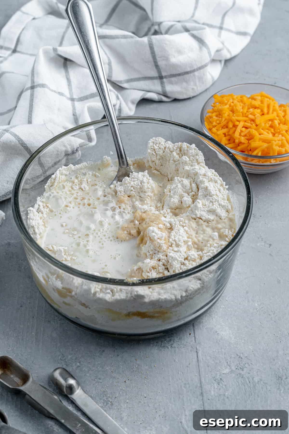 A bowl containing Bisquick mix, milk, and melted butter alongside a separate dish of shredded cheddar cheese.