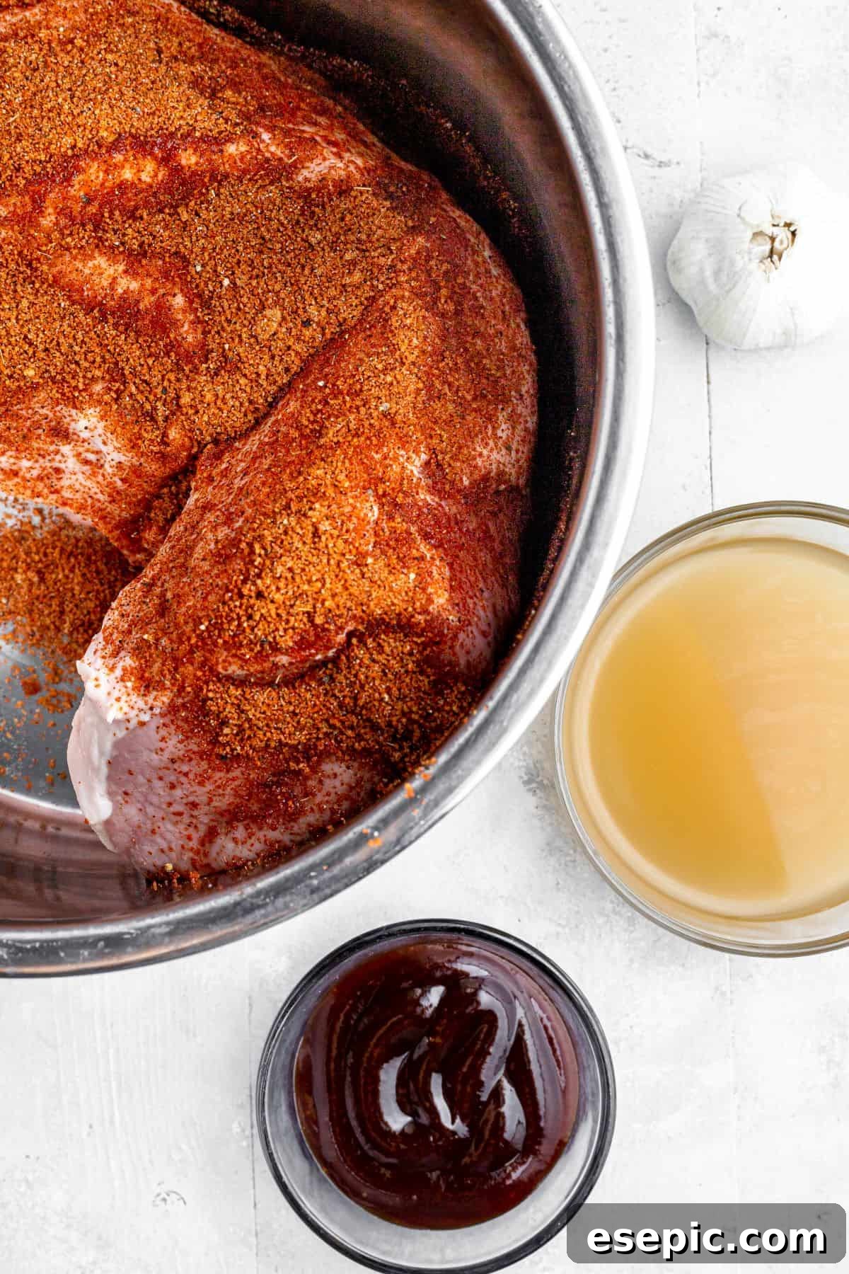 A season-rubbed pork loin in the metal insert of an Instant Pot, with a head of garlic, bowl of broth, and dish of barbecue sauce nearby on the work surface.