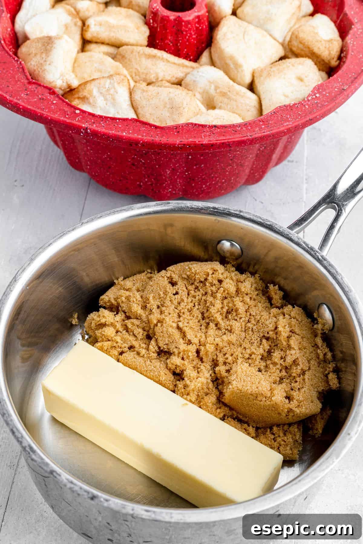 A Bundt pan filled with biscuit dough pieces sits next to a saucepan containing a stick of butter and some brown sugar, ready for the next step.