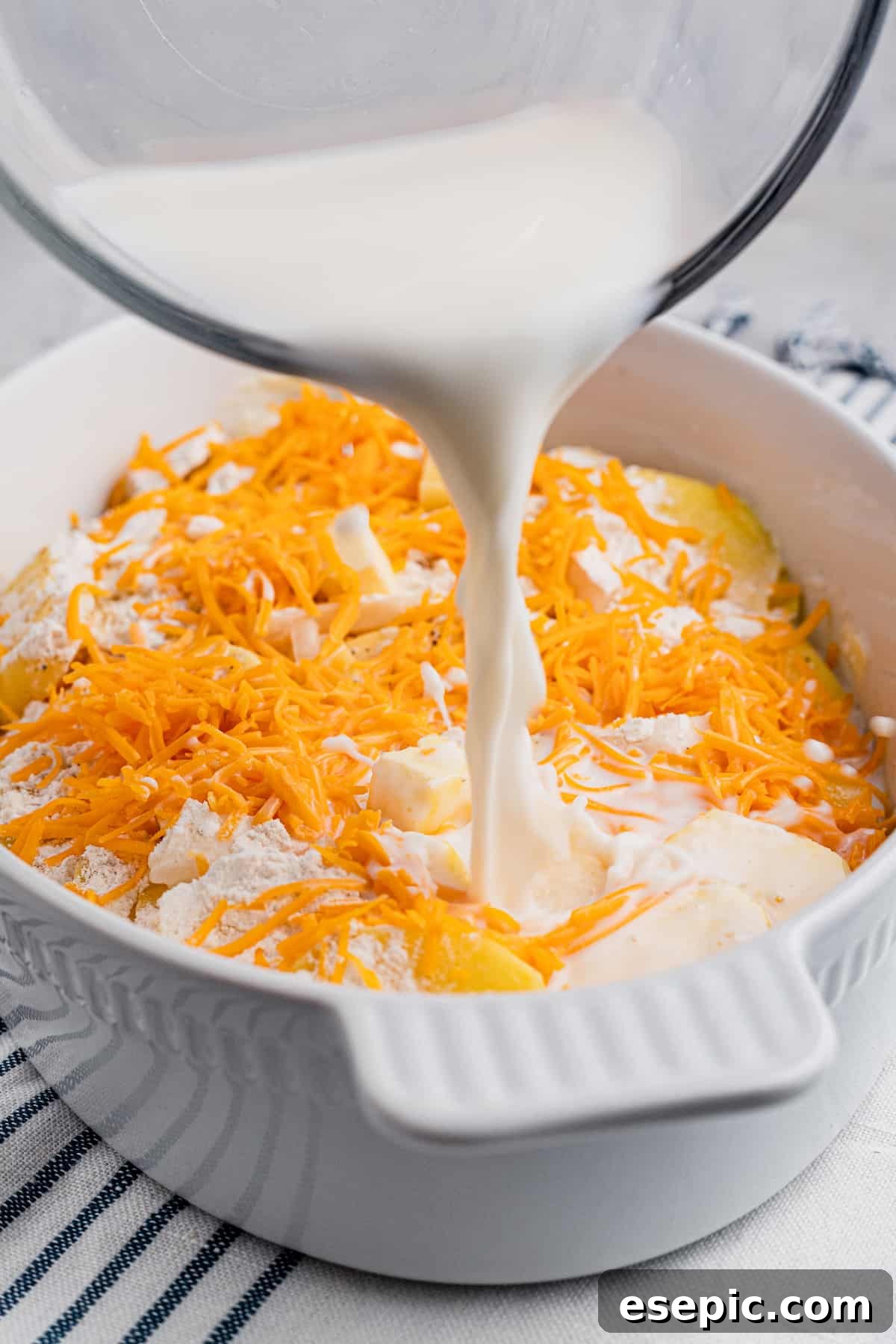 Warm milk being poured over neatly layered sliced potatoes and shredded cheese in a baking dish, ready for baking.