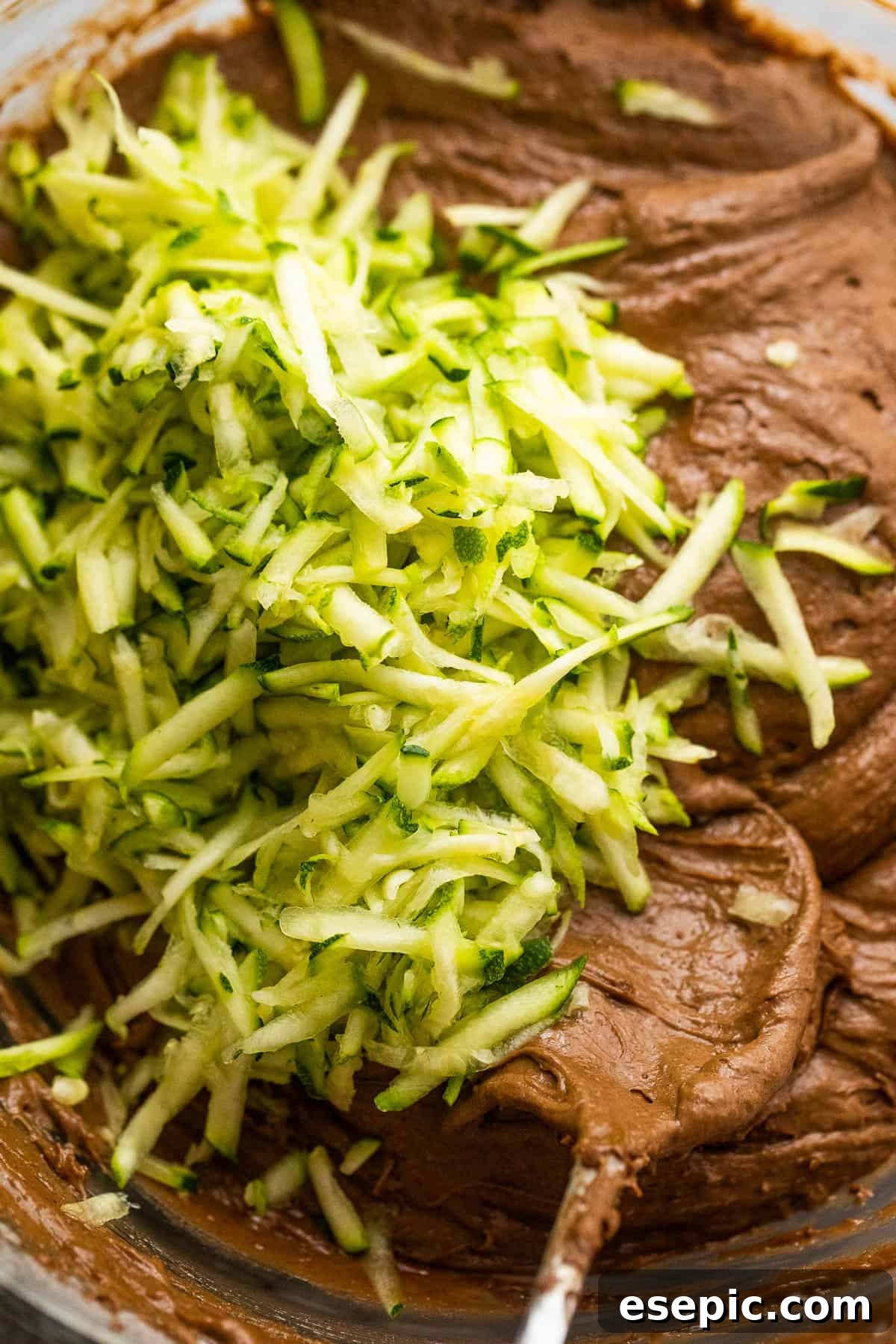 Mixing shredded zucchini into a rich chocolate batter, showing the vibrant green strands blending into the dark mixture.