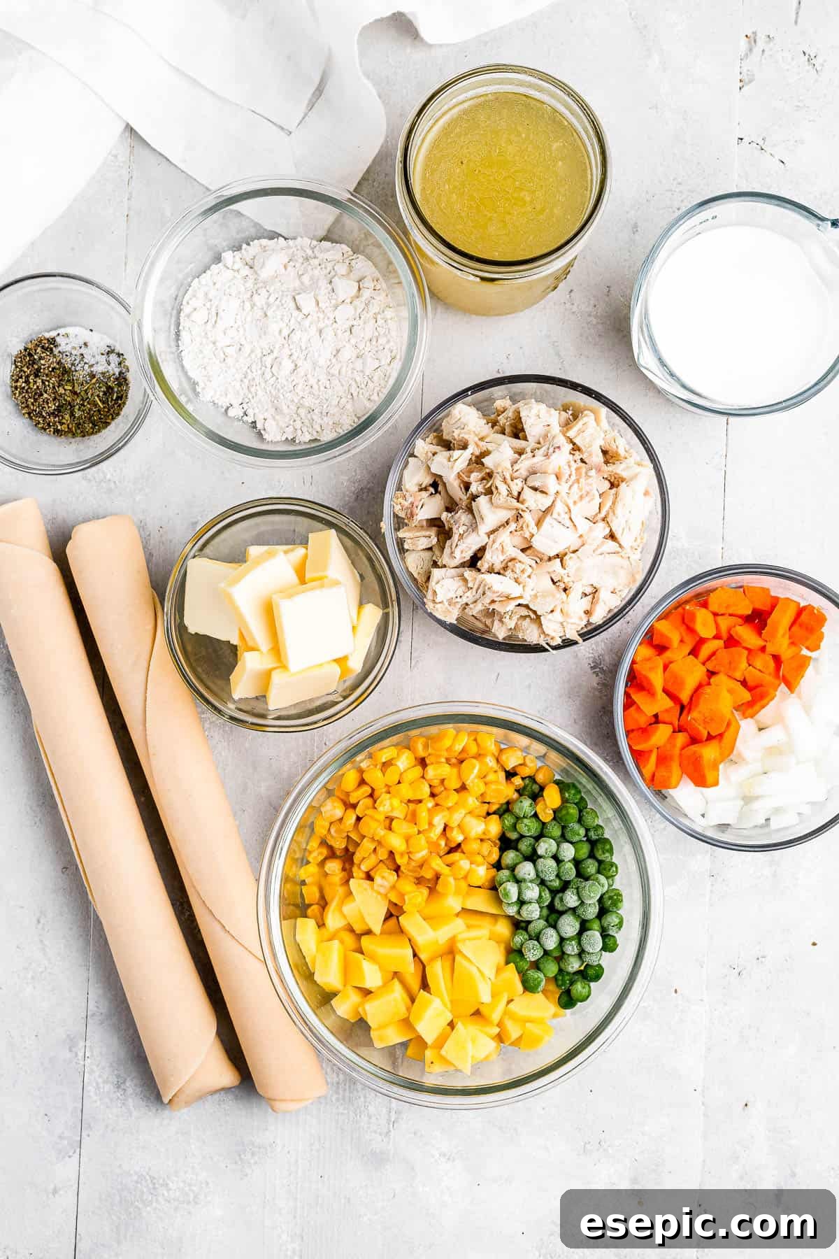 Golden Crust Chicken Delight 3 A vibrant display of fresh ingredients for chicken pot pie, including chicken broth, milk, chopped carrots and onions, a bowl of peas, corn, and diced potatoes, butter, various seasonings, and flour, arranged neatly on a surface.