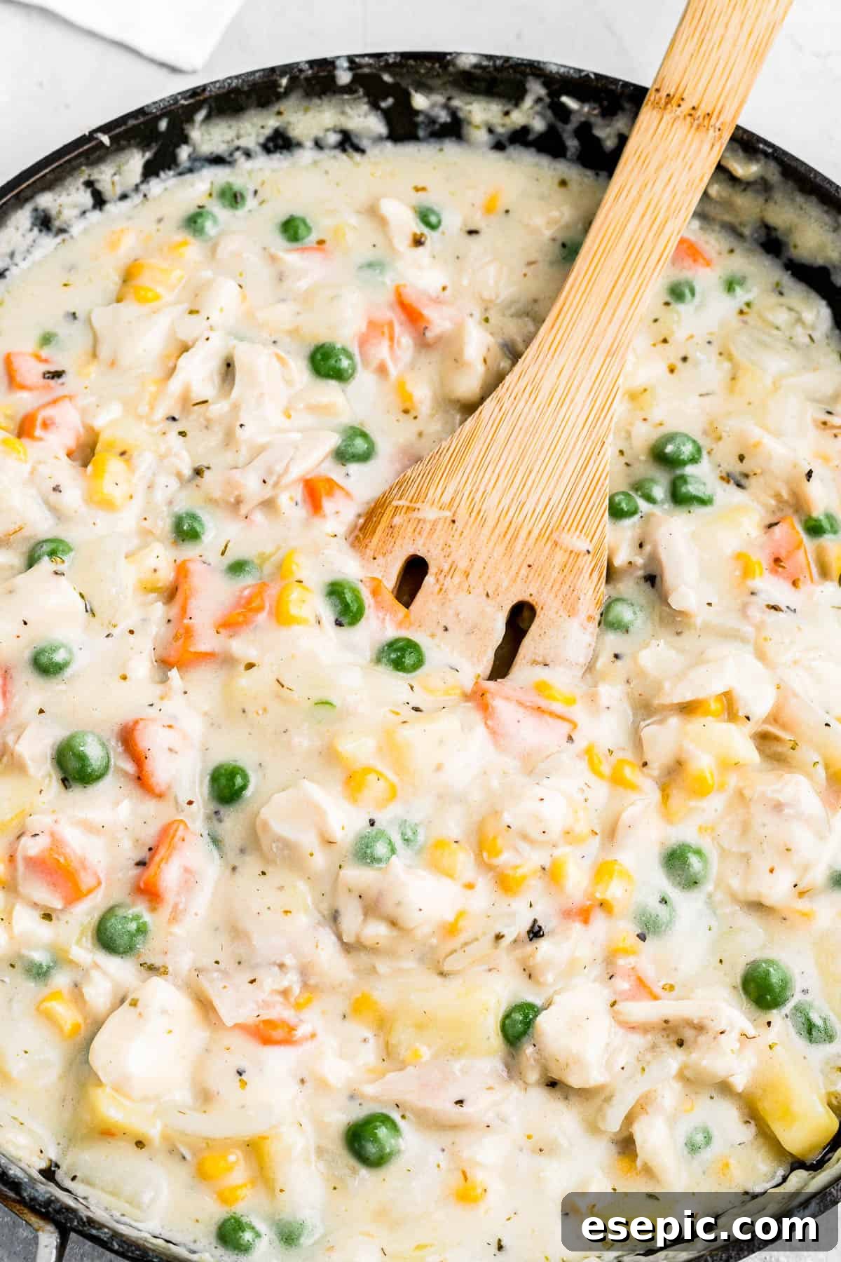 Golden Crust Chicken Delight 4 A wooden spoon gently stirs the rich, creamy chicken pot pie filling in a skillet, showcasing a delightful blend of tender chicken, vibrant carrots, plump peas, sweet corn, and perfectly diced potatoes.