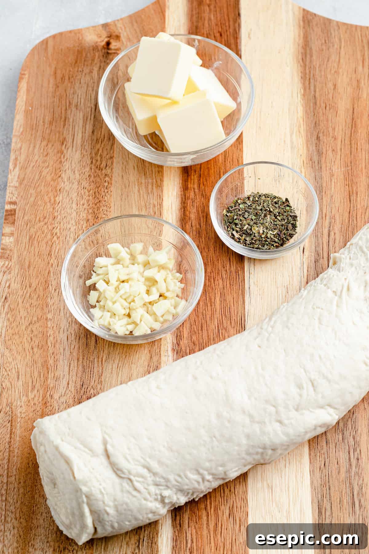 Clockwise from top: butter, Italian seasoning, a roll of pizza dough, a dish of minced garlic.