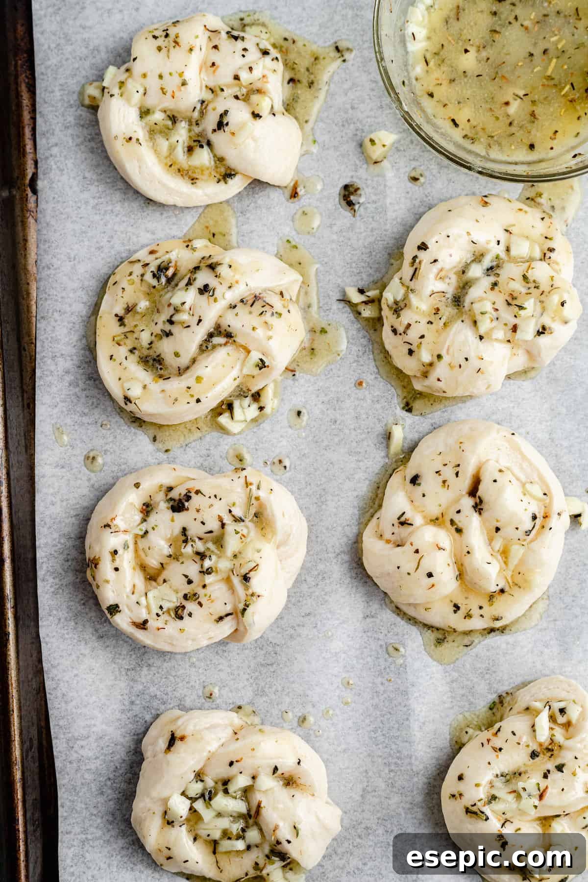 Unbaked garlic knots, brushed with garlic-and-herb-butter, on a parchment-lined tray.