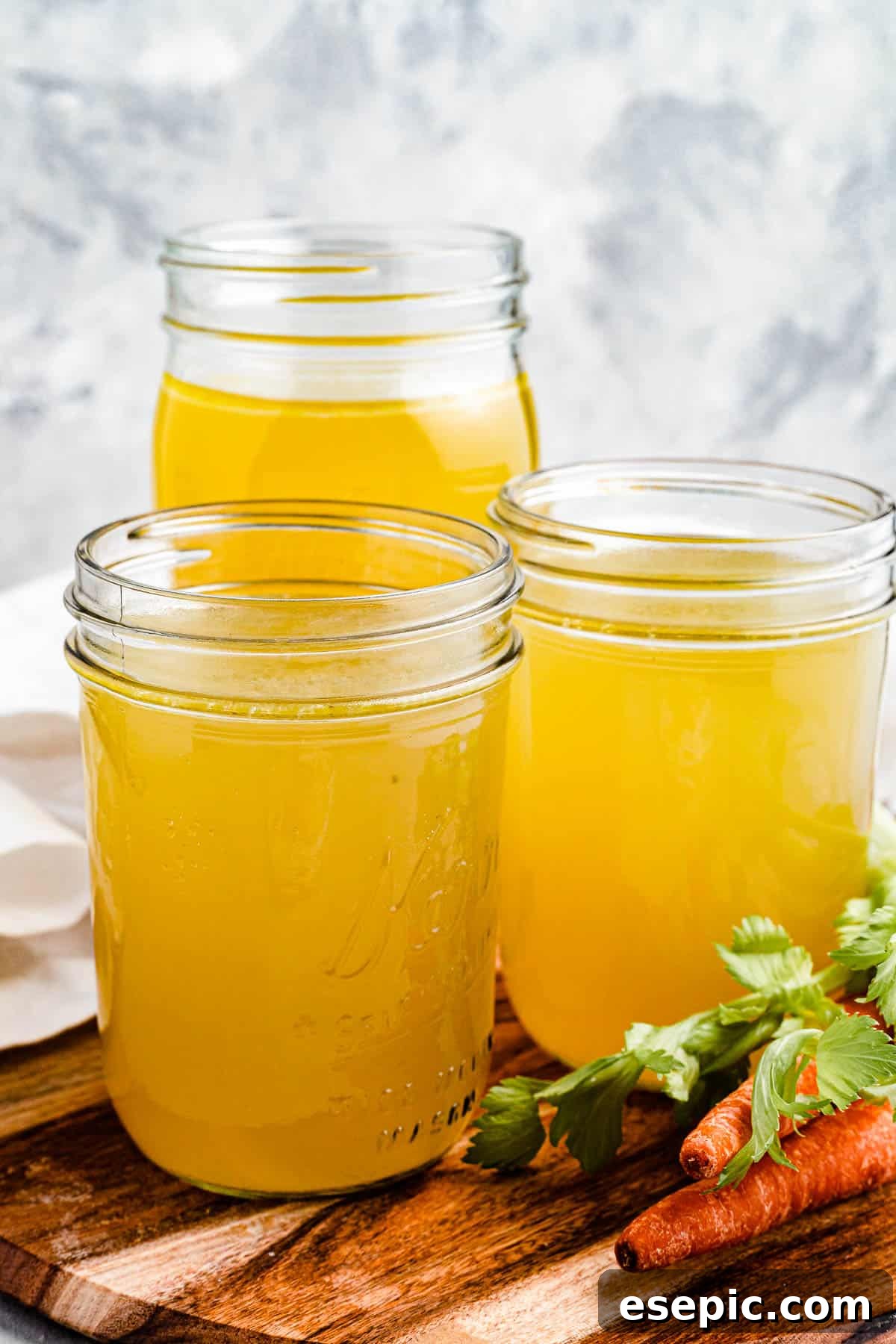Three jars of homemade chicken broth on a rustic cutting board, with fresh raw carrots nearby, symbolizing wholesome ingredients.